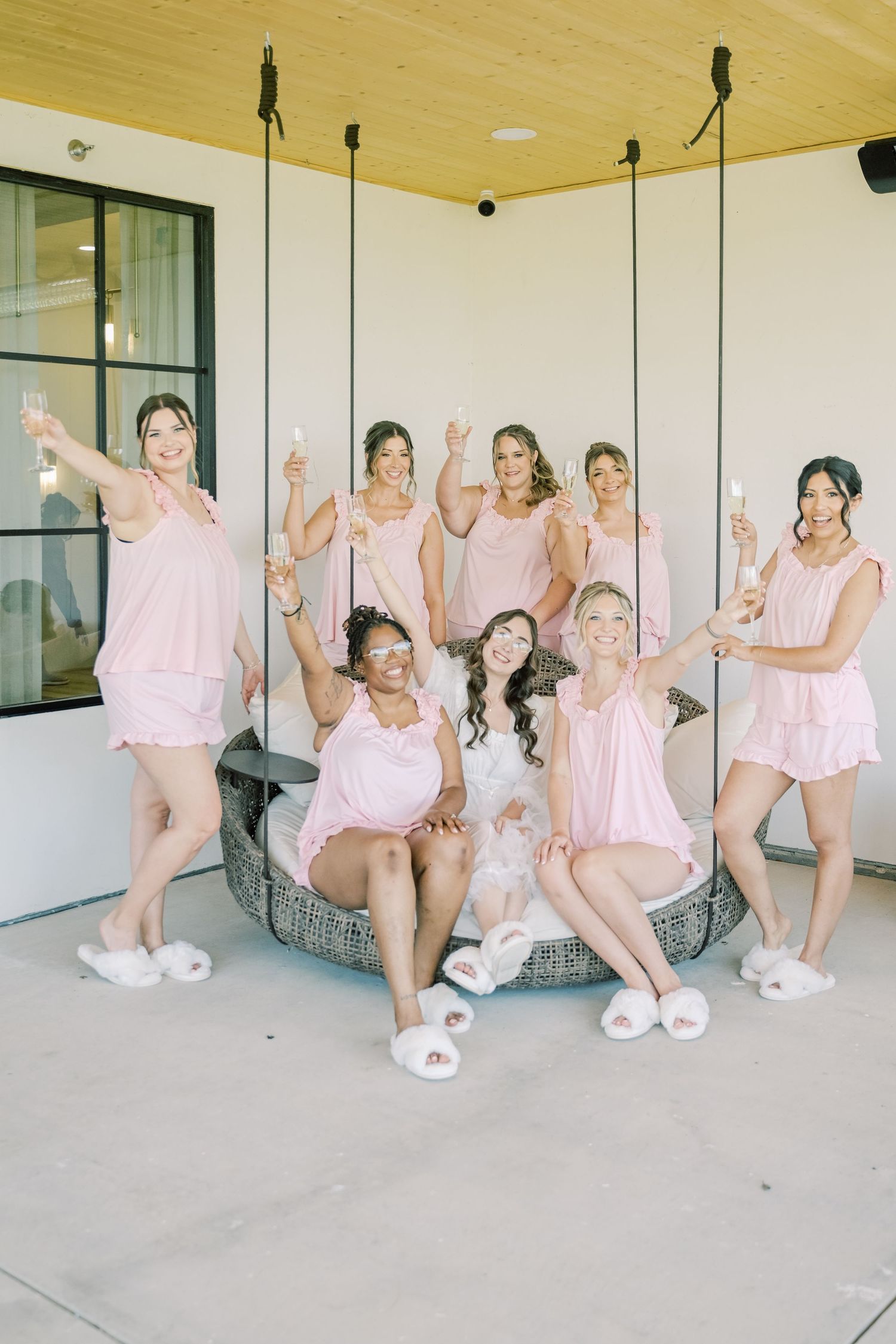Bridesmaids in matching pink pajamas and white fluffy slippers toast champagne on a hanging swing seat.