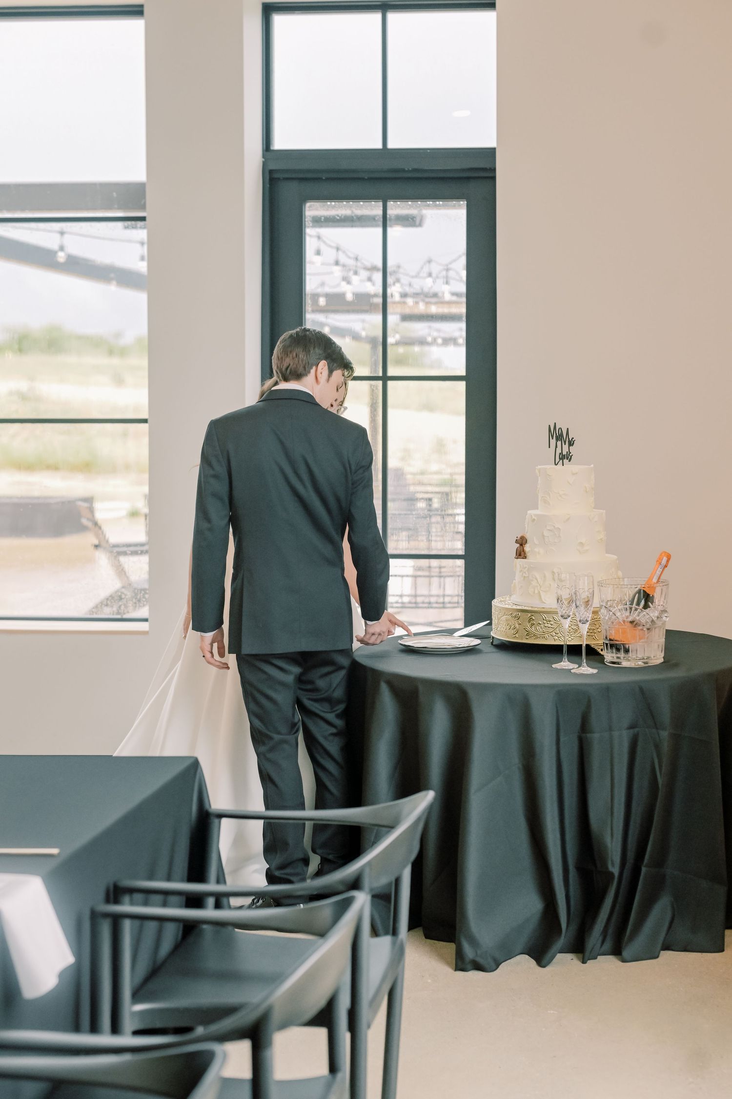 Groom in black suit standing by window near wedding cake table at reception venue.
