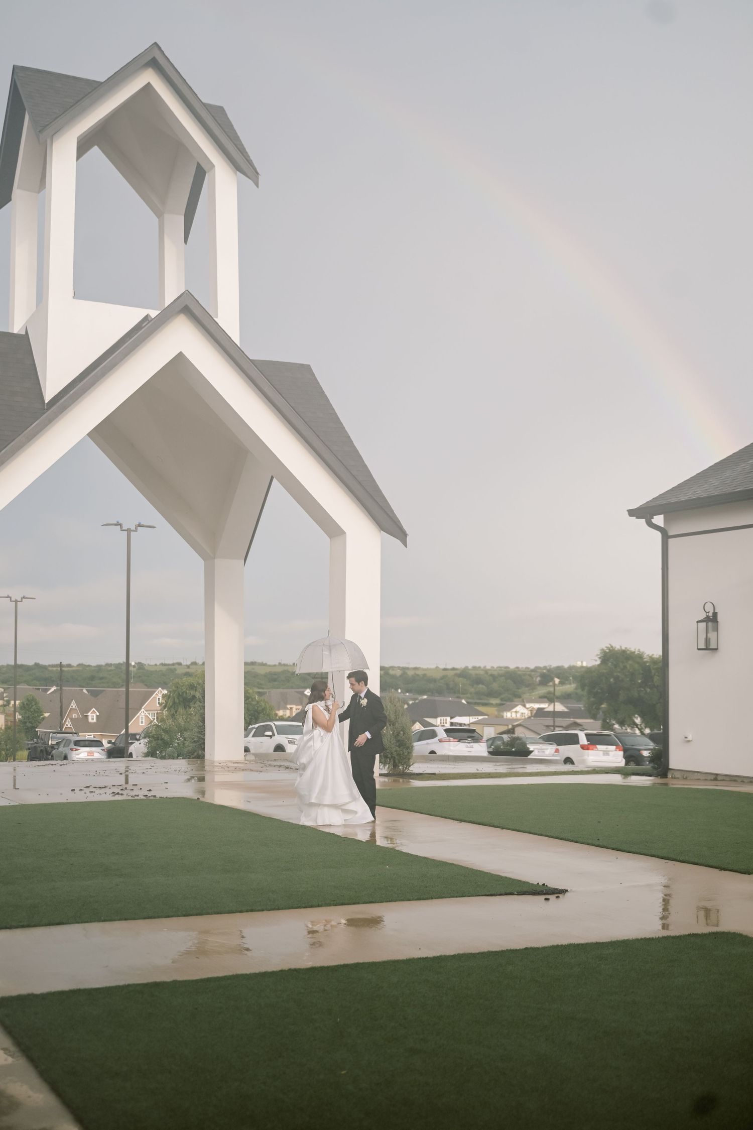 Bride and groom sharing a romantic moment under a stunning rainbow outside a white wedding venue in the rain.