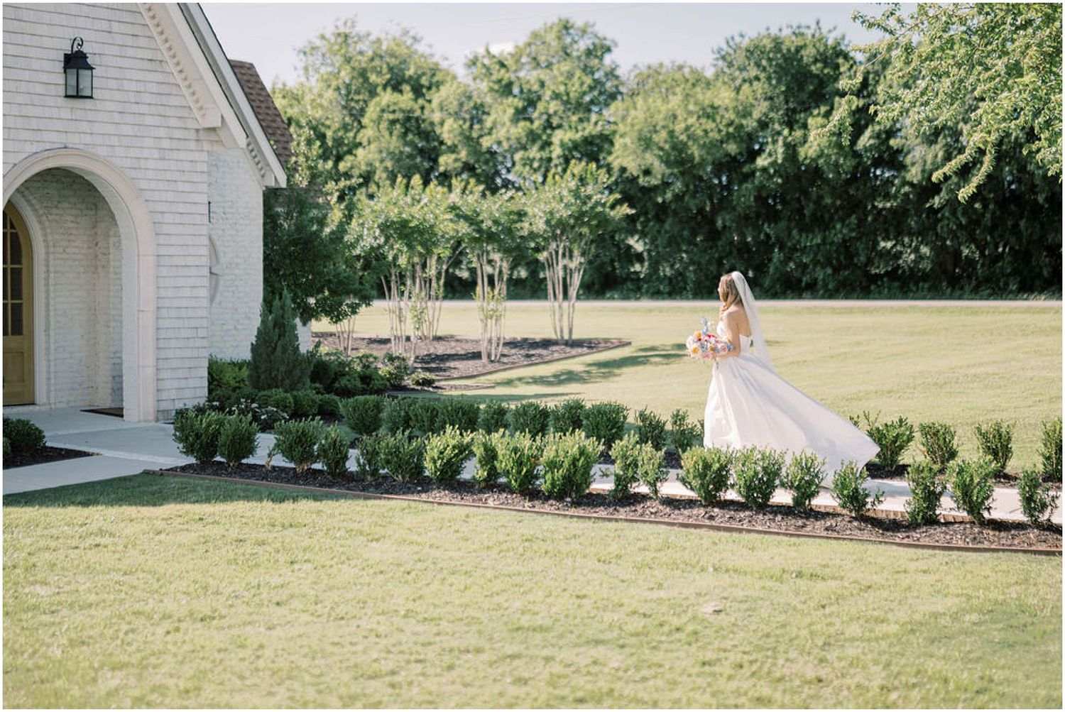 Bride in a flowing white gown walks along a manicured garden path beside a white chapel on a sunny day.