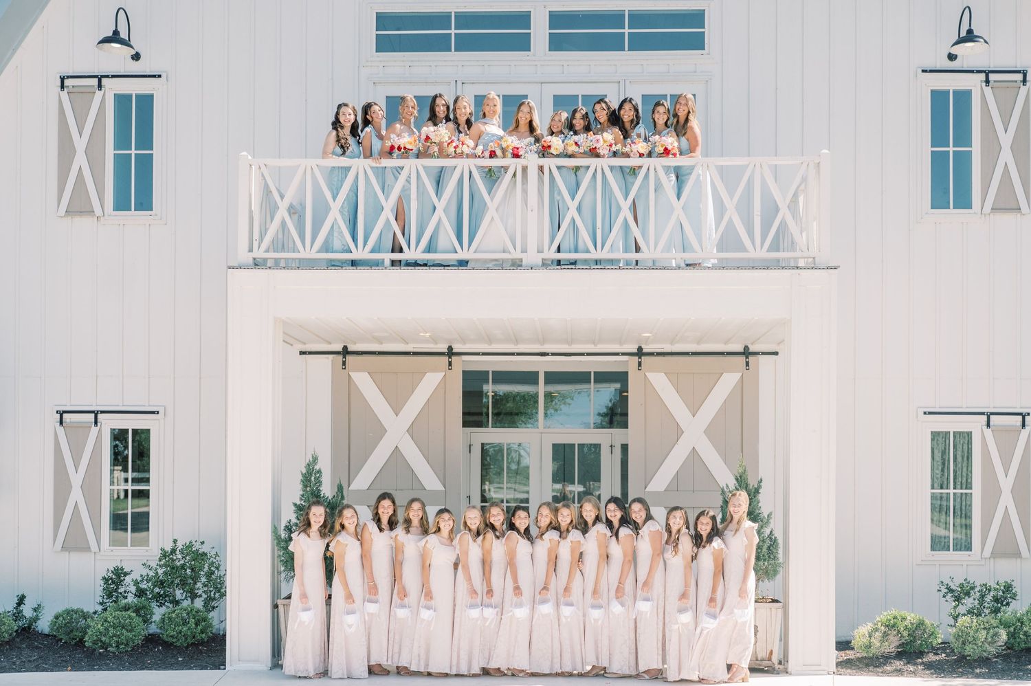 Large bridal party poses in white gowns outside a white barn venue, with more bridesmaids waving from the balcony above.