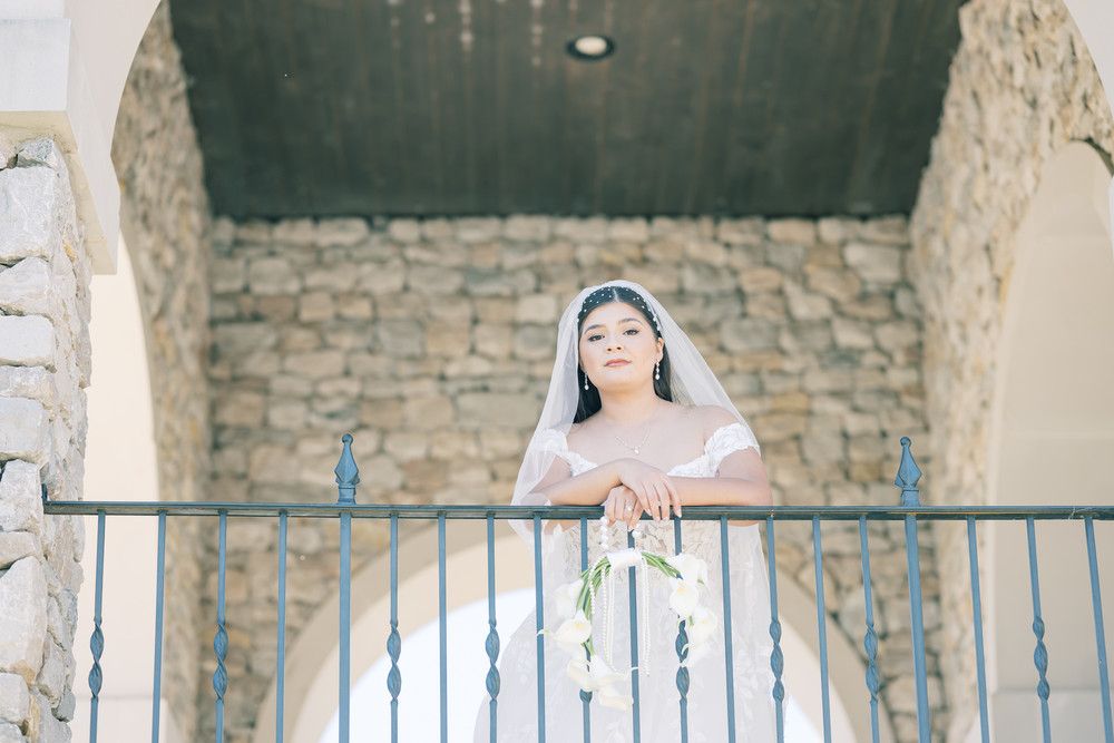 A bride in a white lace gown poses elegantly against a rustic stone wall with arched windows.