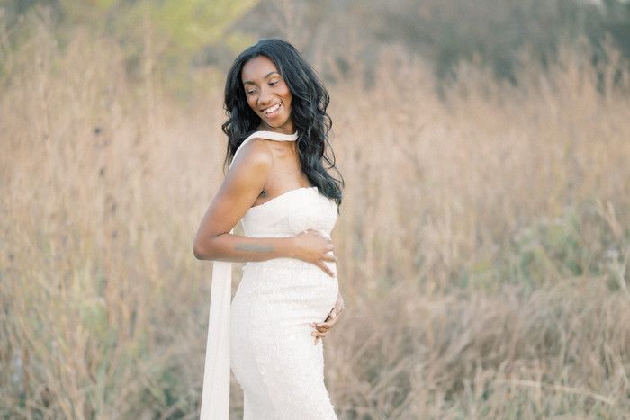 Maternity portrait of an expectant mother in a white dress posing in a golden field at sunset.