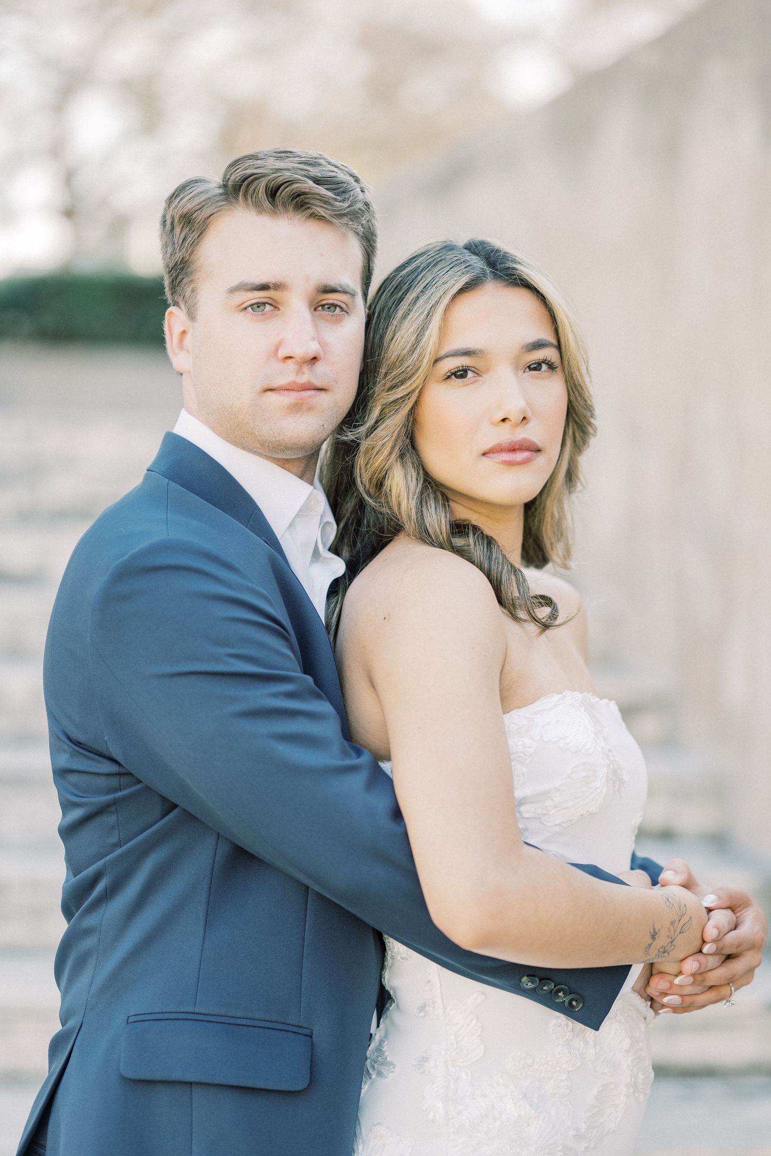 A romantic wedding couple embraces in soft natural light while wearing a navy suit and white lace gown.