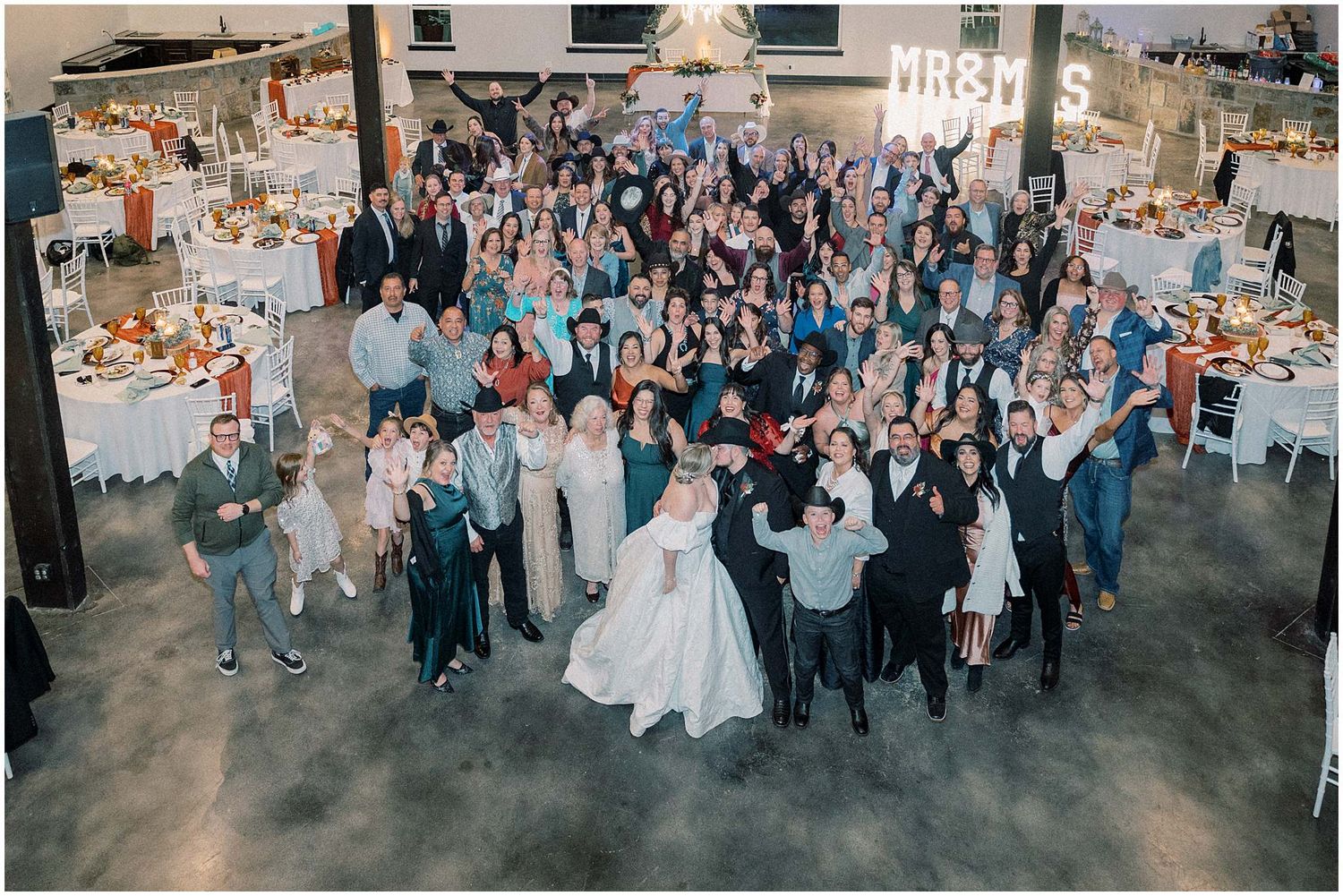 Large group of wedding guests gathered together for a panoramic photo in an indoor venue.