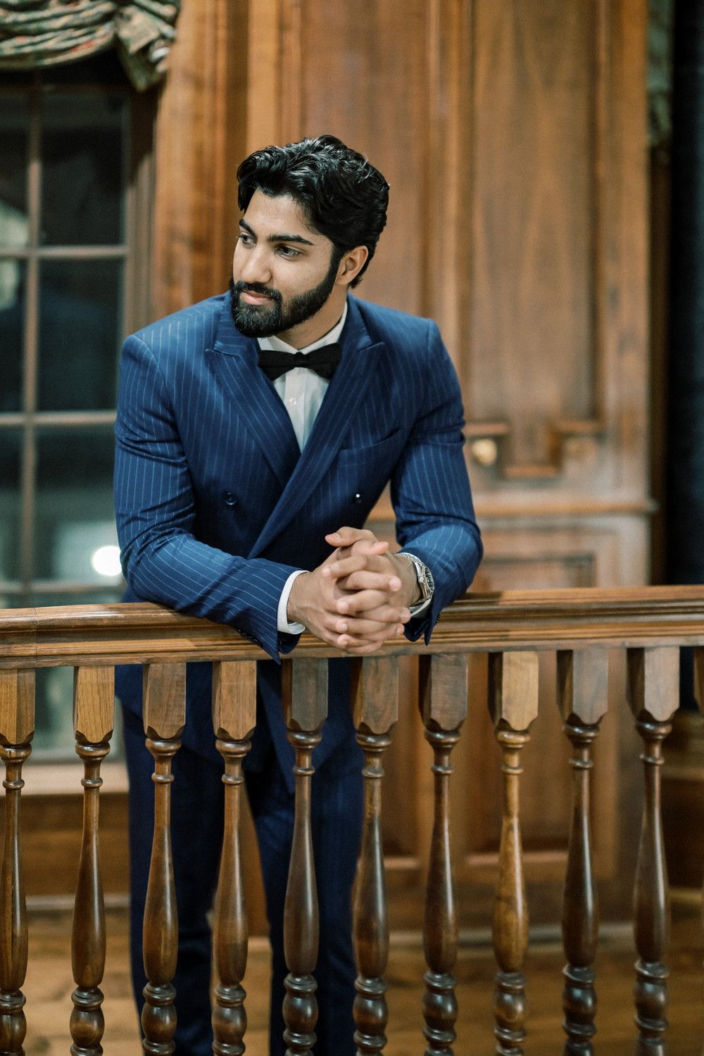 Man in navy suit thoughtfully gazes while leaning on ornate wooden railing.
