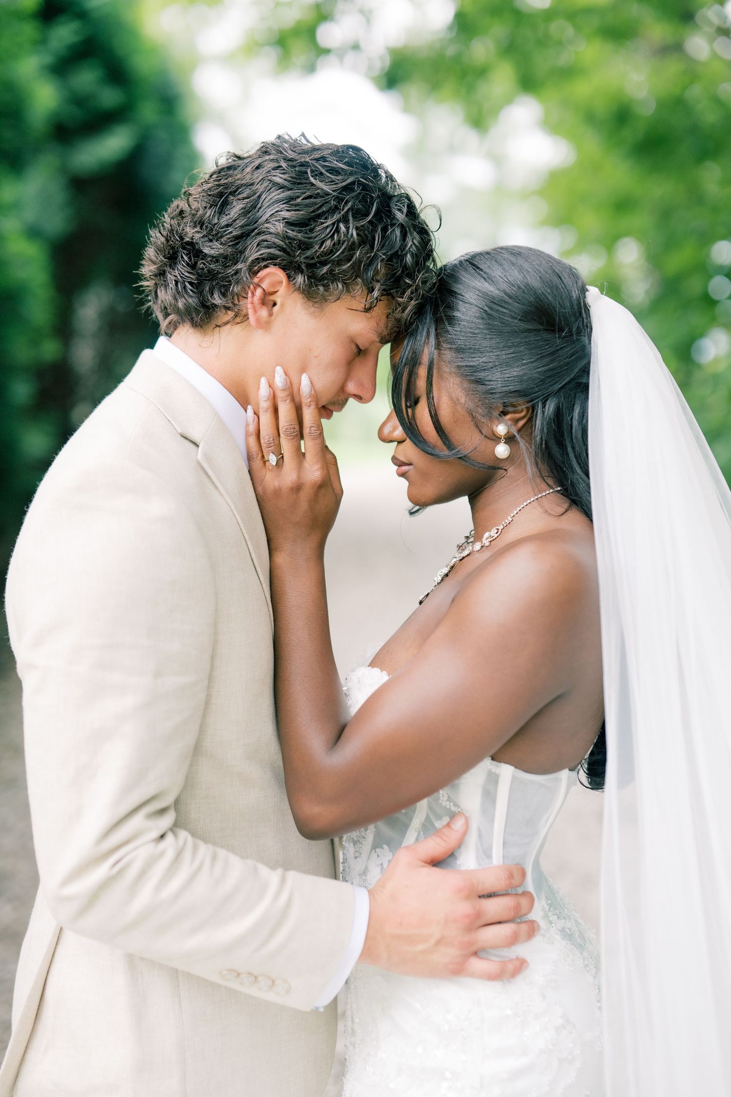 Interracial couple sharing an intimate wedding moment, groom in beige suit and bride in lace gown with veil outdoors.