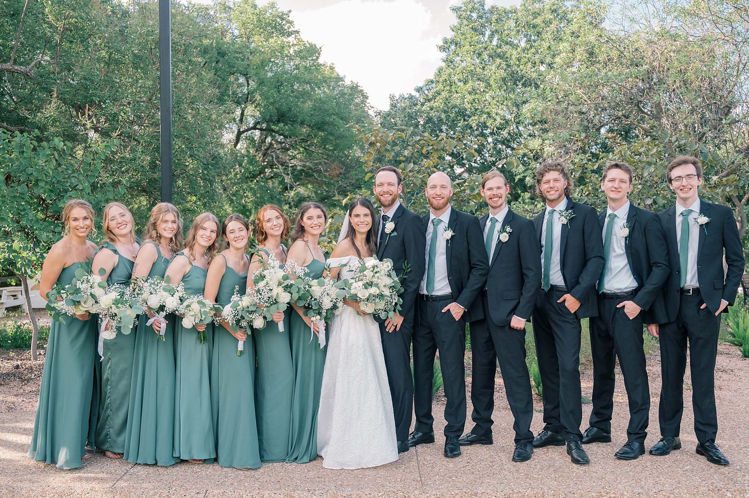 Wedding party members in sage green dresses and black suits gather for formal photos outdoors.