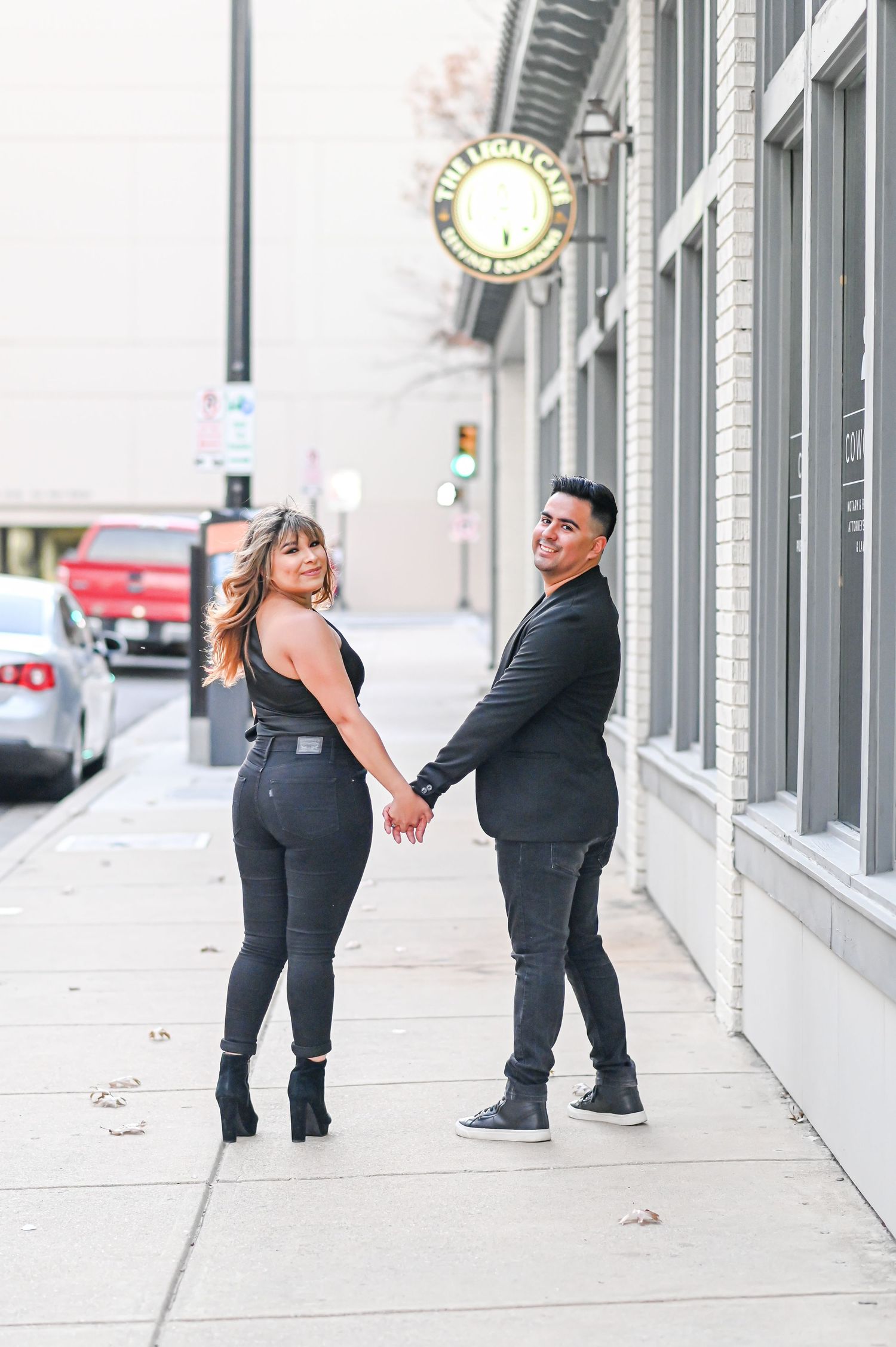 Couple in black outfits holding hands and posing on a city sidewalk near a clock.