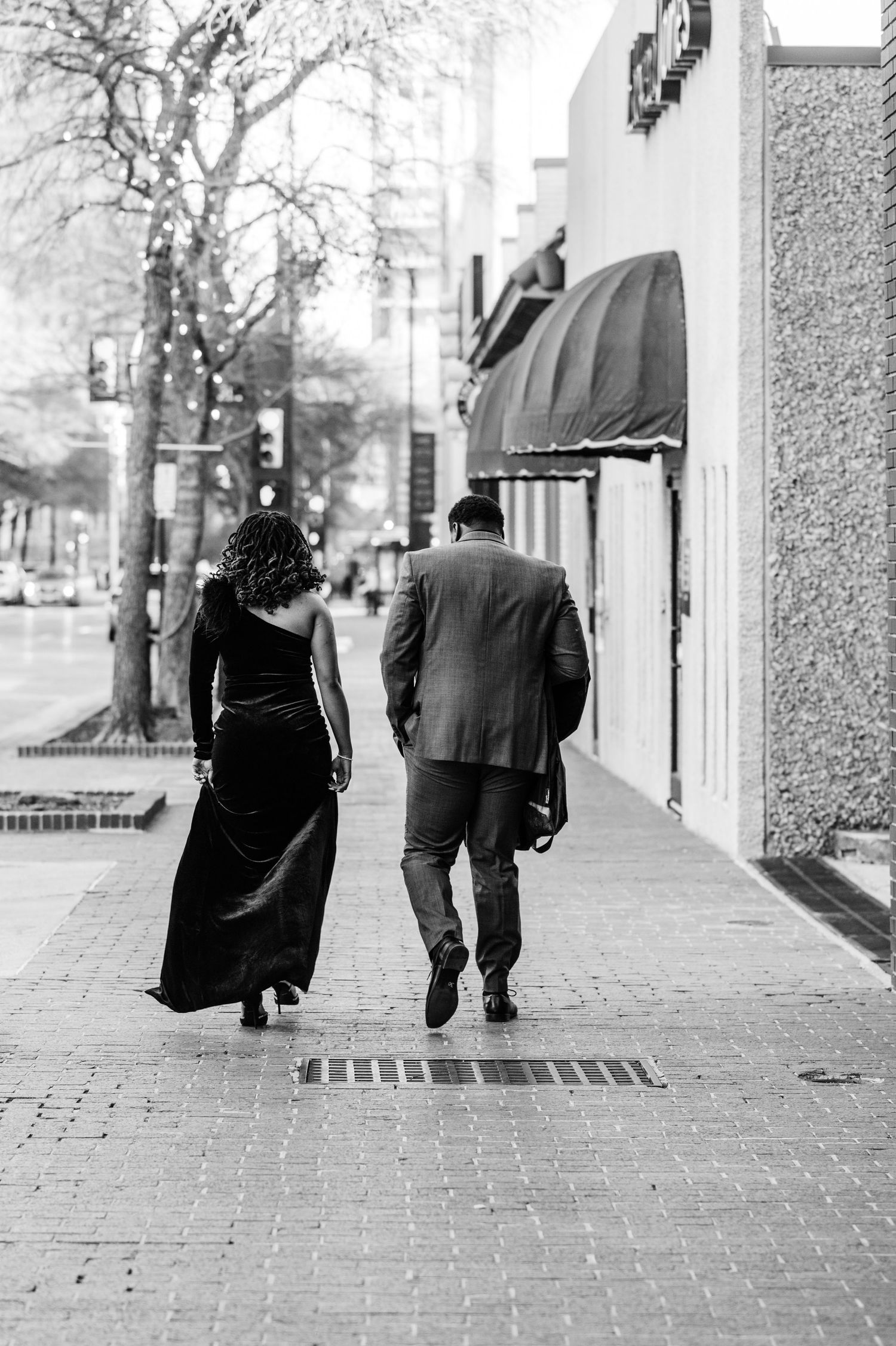 Black and white photo of a couple walking on a city sidewalk, woman in elegant gown and man in suit.