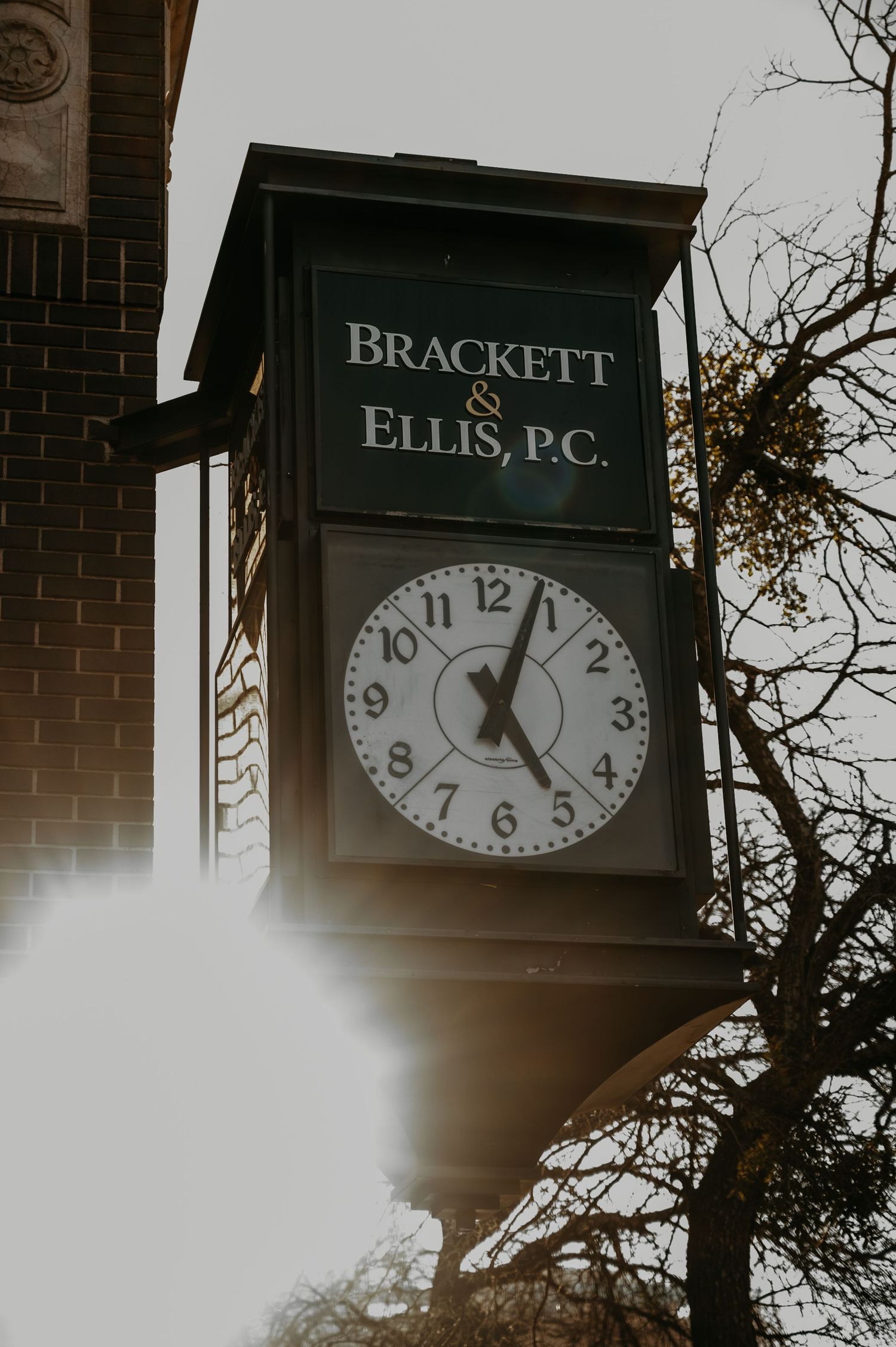 Brackett & Ellis P.C. law firm outdoor clock sign mounted on brick building with bare trees in background.