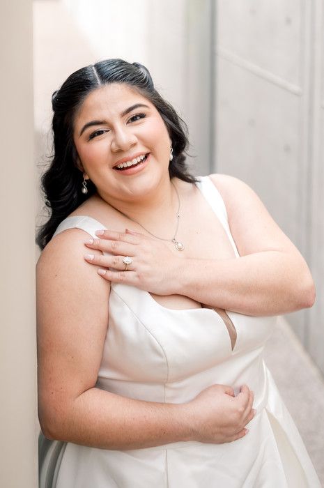 Smiling bride in white dress with diamond necklace and ring, leaning against a wall in a joyful portrait.