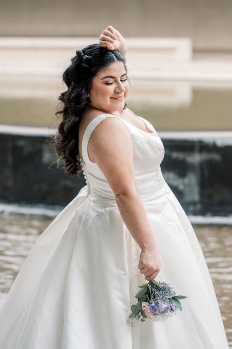 Bride in white ball gown holding bouquet of flowers, smiling with hand in hair near fountain.