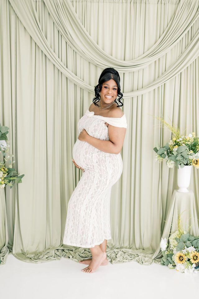 Pregnant woman in white lace off-shoulder gown poses against sage green draped backdrop with floral arrangements.