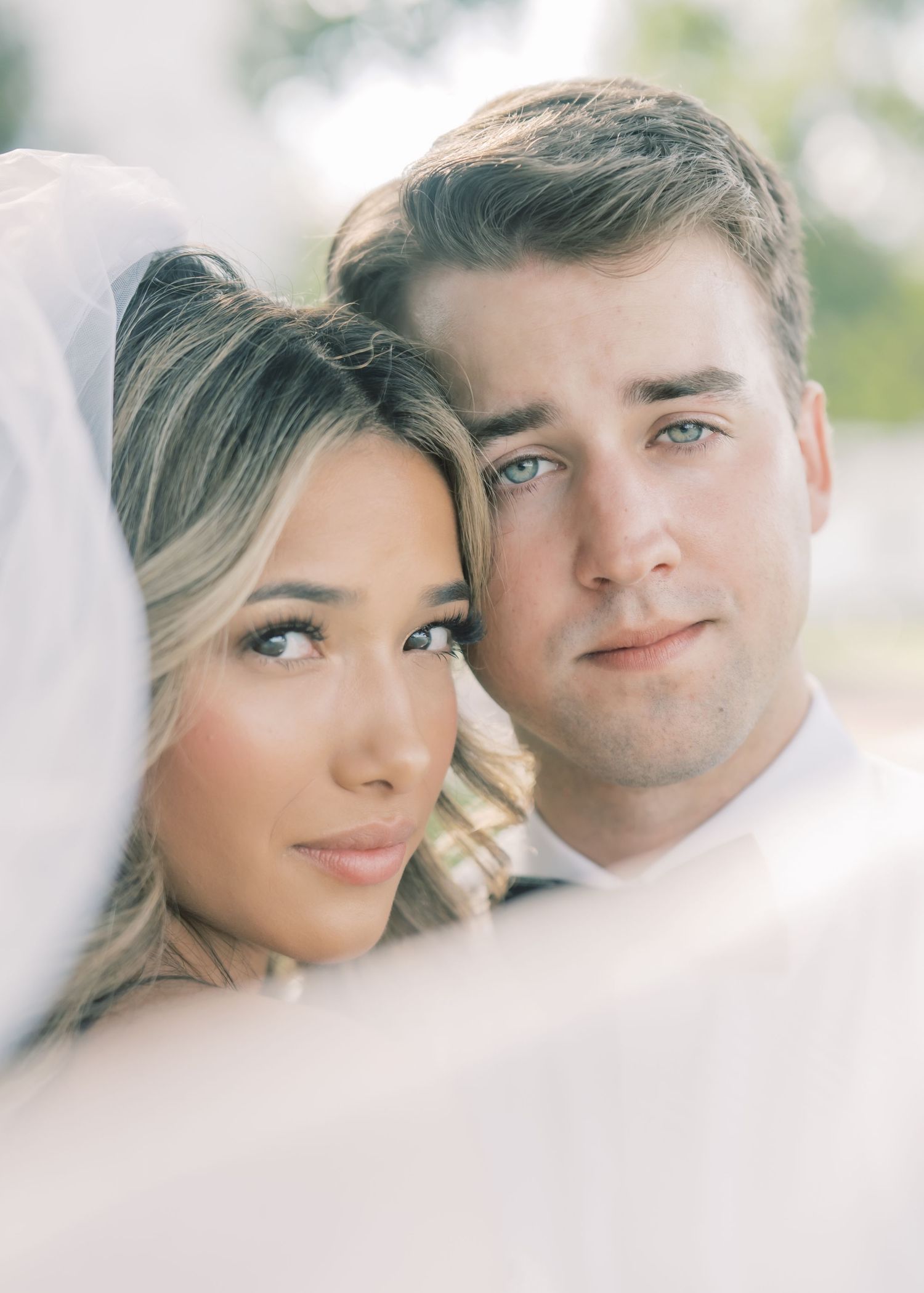 A bride and groom pose closely together under a white veil in a romantic outdoor wedding portrait.