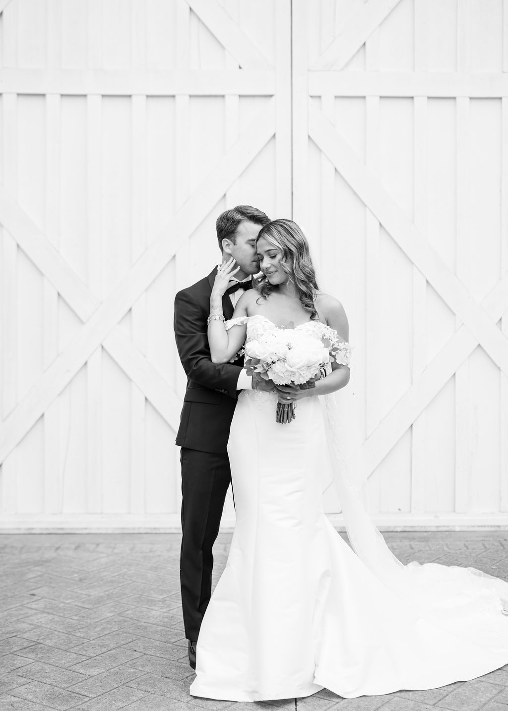Black and white photo of groom kissing bride holding bouquet outside white barn doors.
