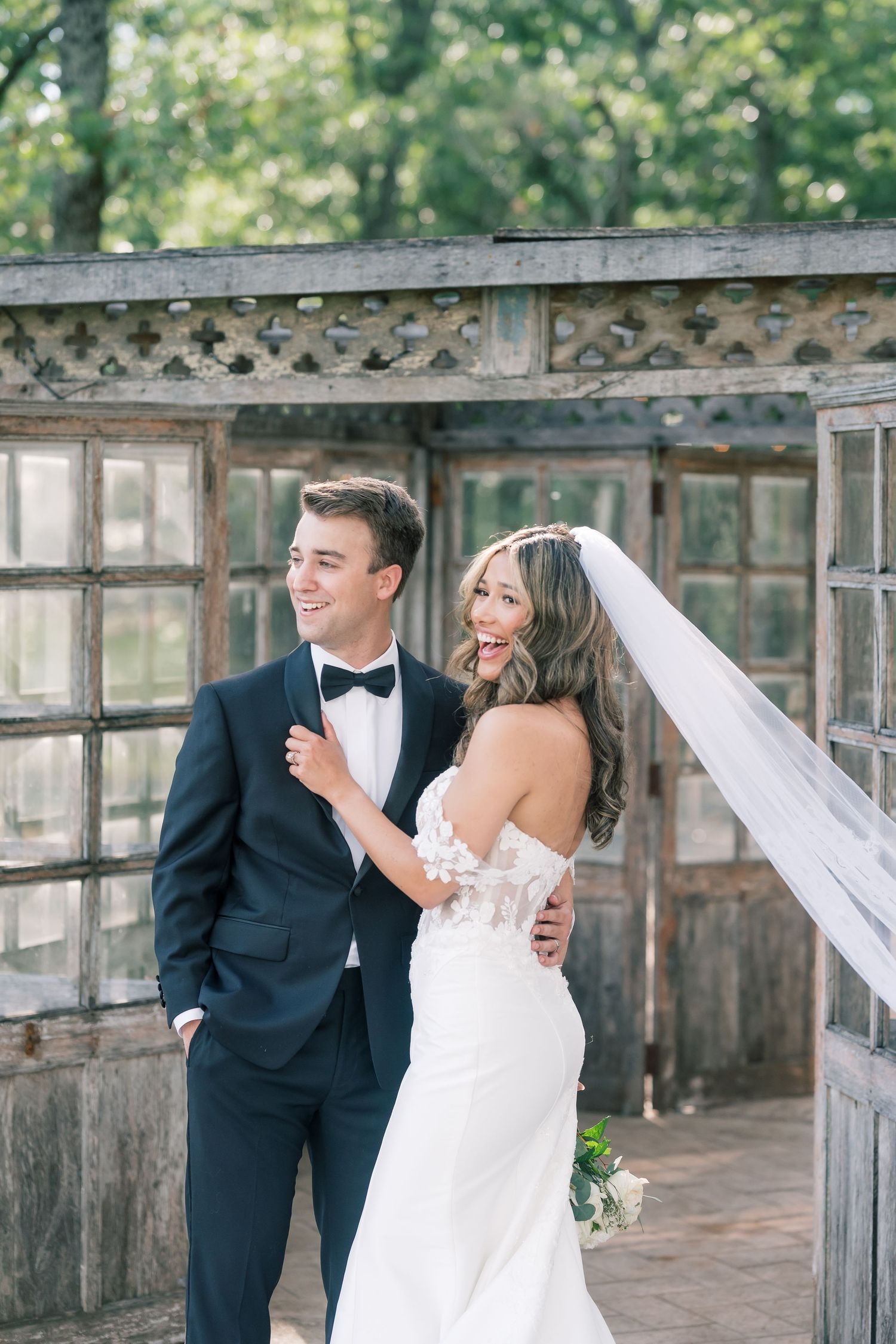 Smiling bride and groom pose together outdoors in wedding attire near a vintage wooden greenhouse.