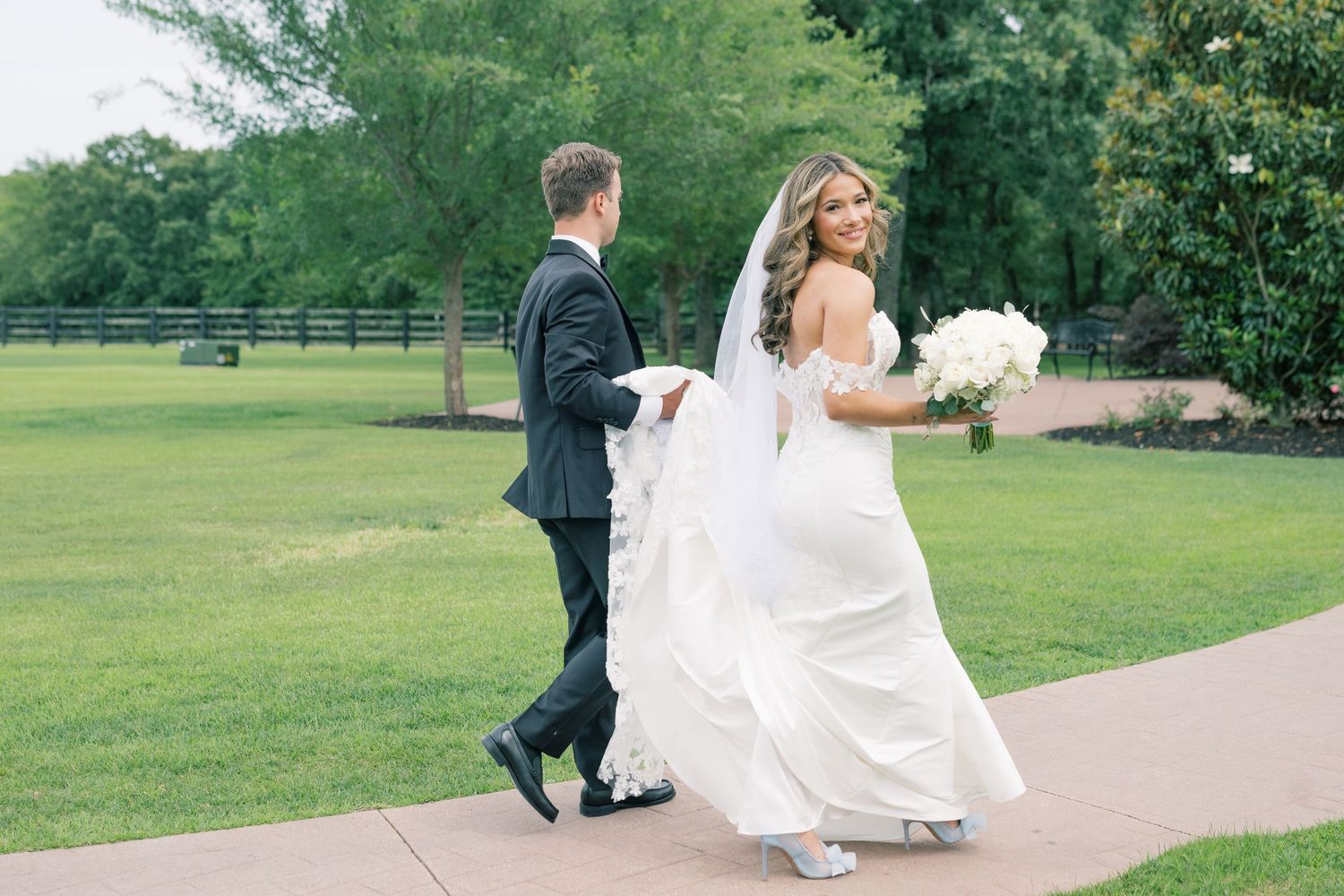 Bride in white mermaid gown holding bouquet walks with groom along pathway at outdoor wedding venue.
