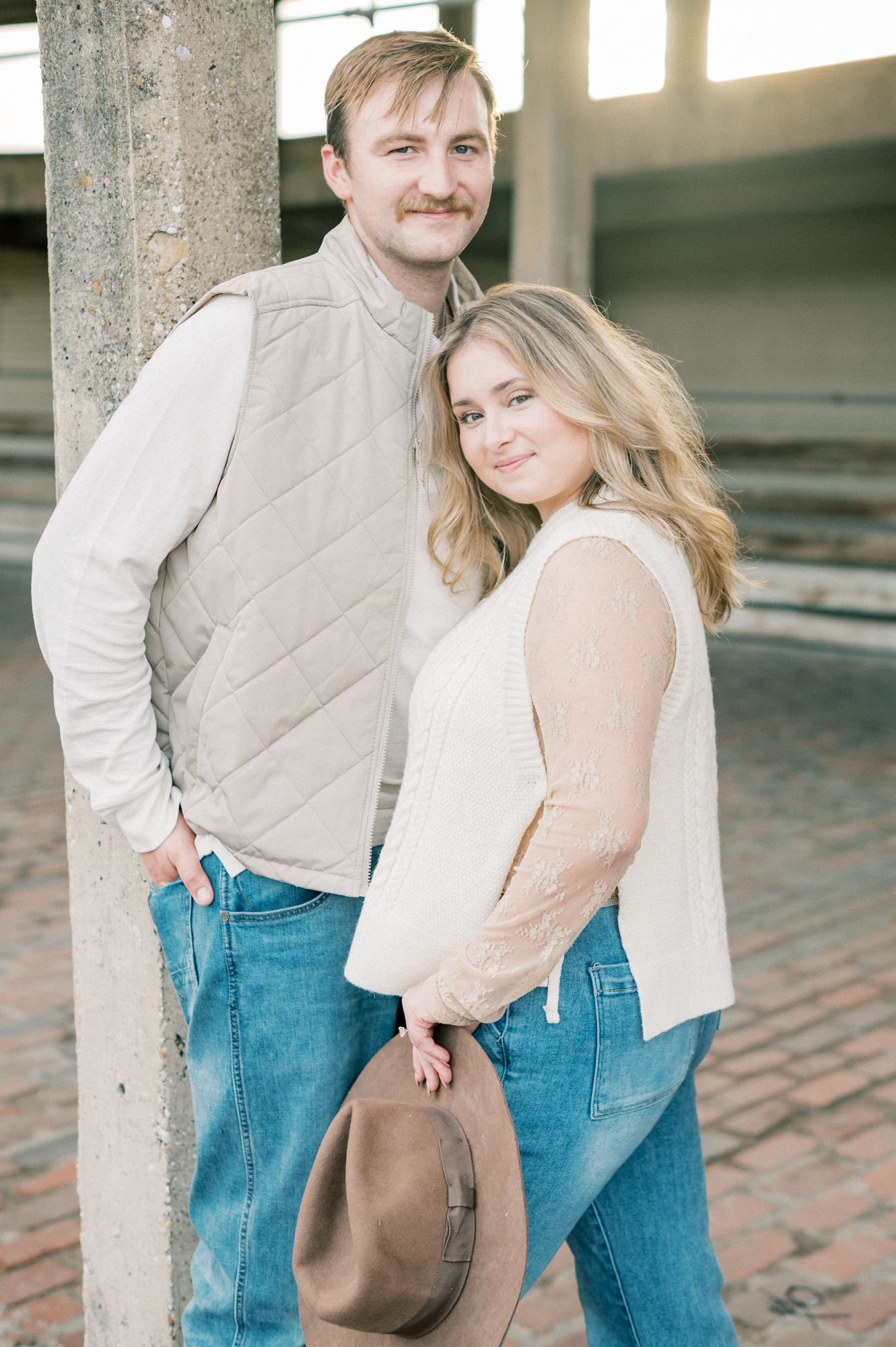 A couple in blue jeans and light tops embrace while standing between concrete pillars.