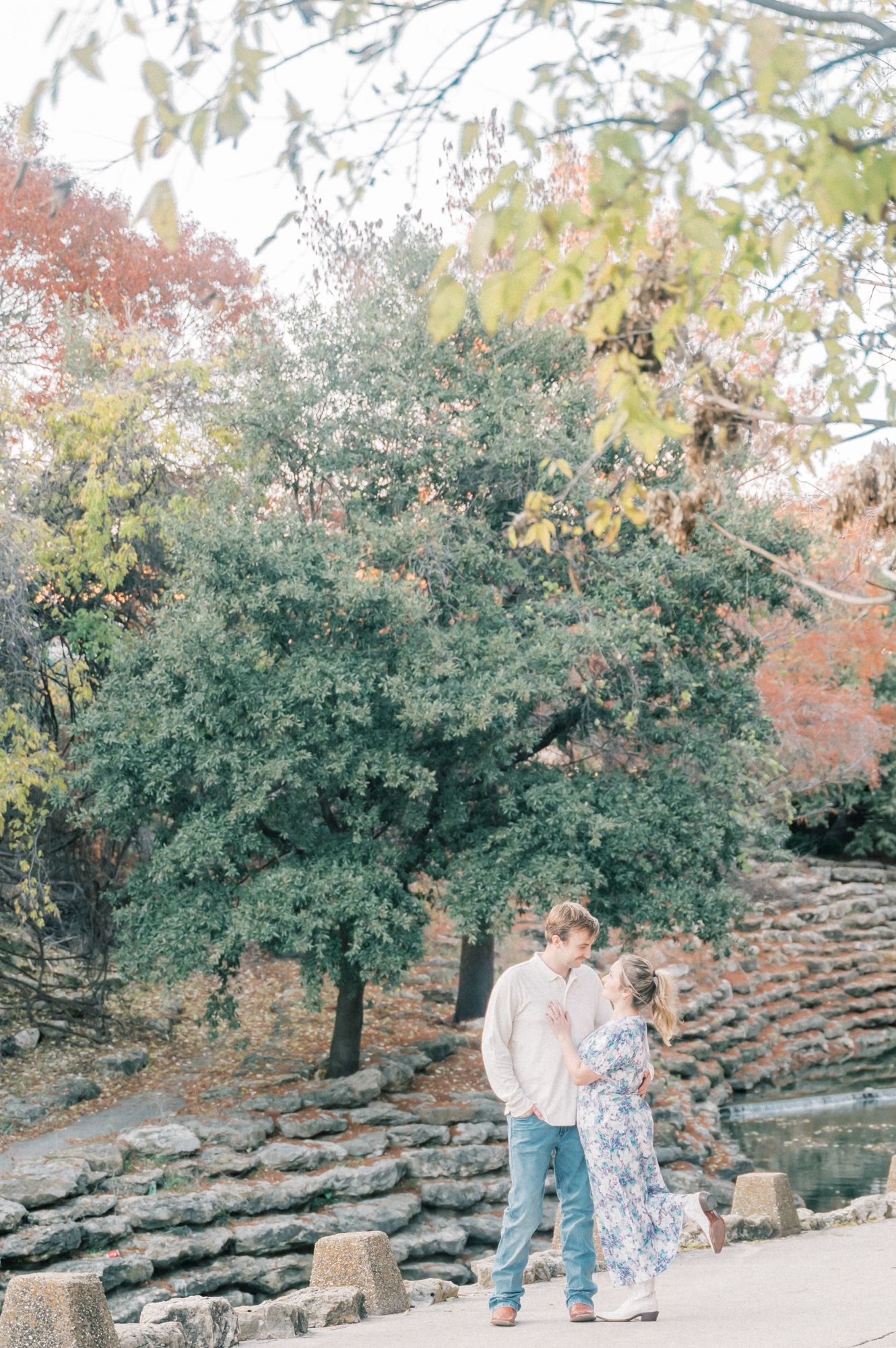 A couple walks together in a romantic autumn setting with stone walls and fall foliage in the background.