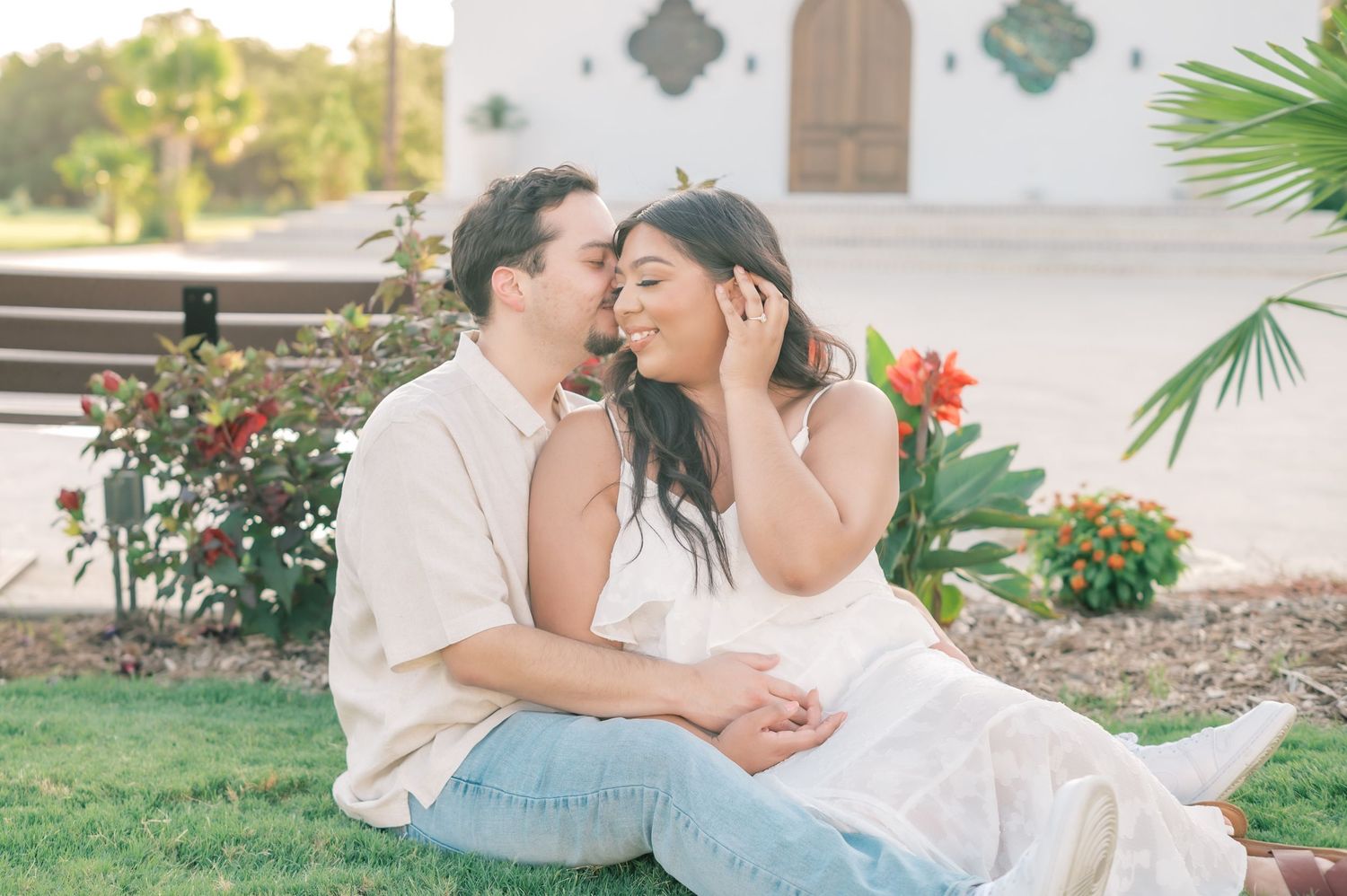 A romantic couple shares tender moments in a garden setting with palm trees and flowers in the background.