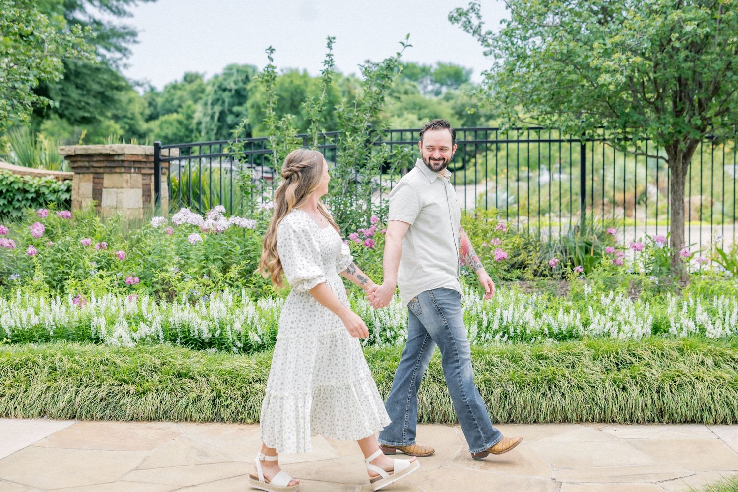 Couple holding hands walking through a lush garden with blooming flowers and iron gate backdrop.