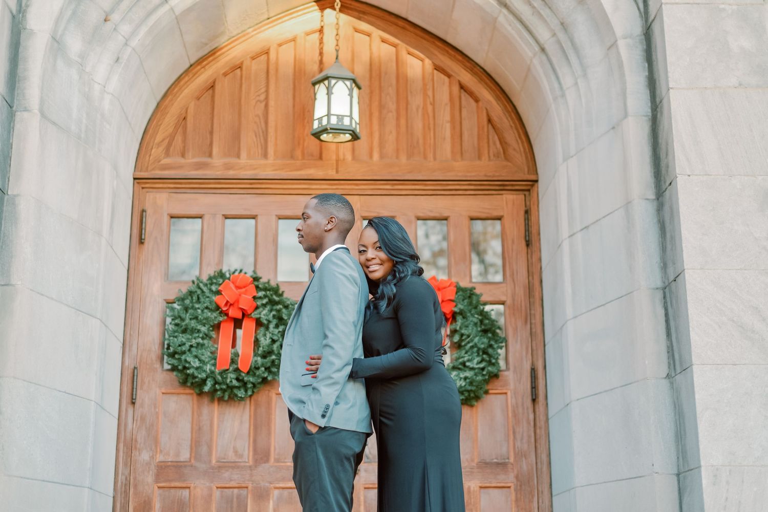 Couple poses back-to-back before wooden arched doors adorned with Christmas wreaths at a stone building.