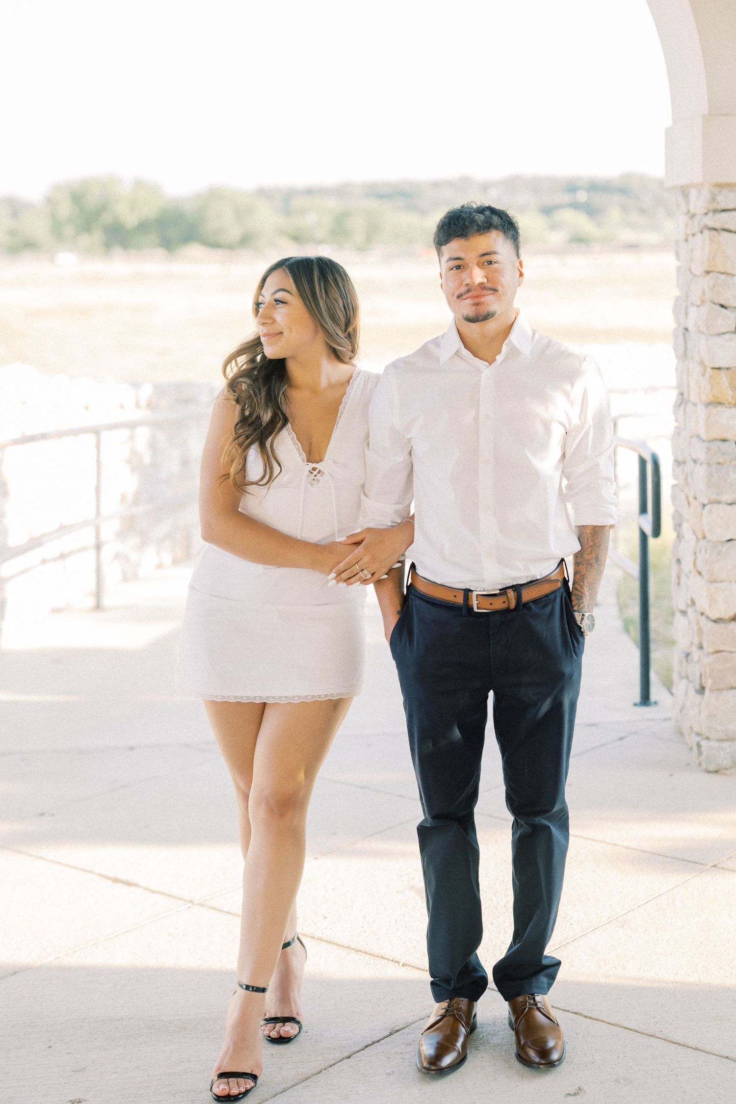 A couple in white outfits poses outdoors, woman in mini dress with heels, man in white shirt and navy pants.
