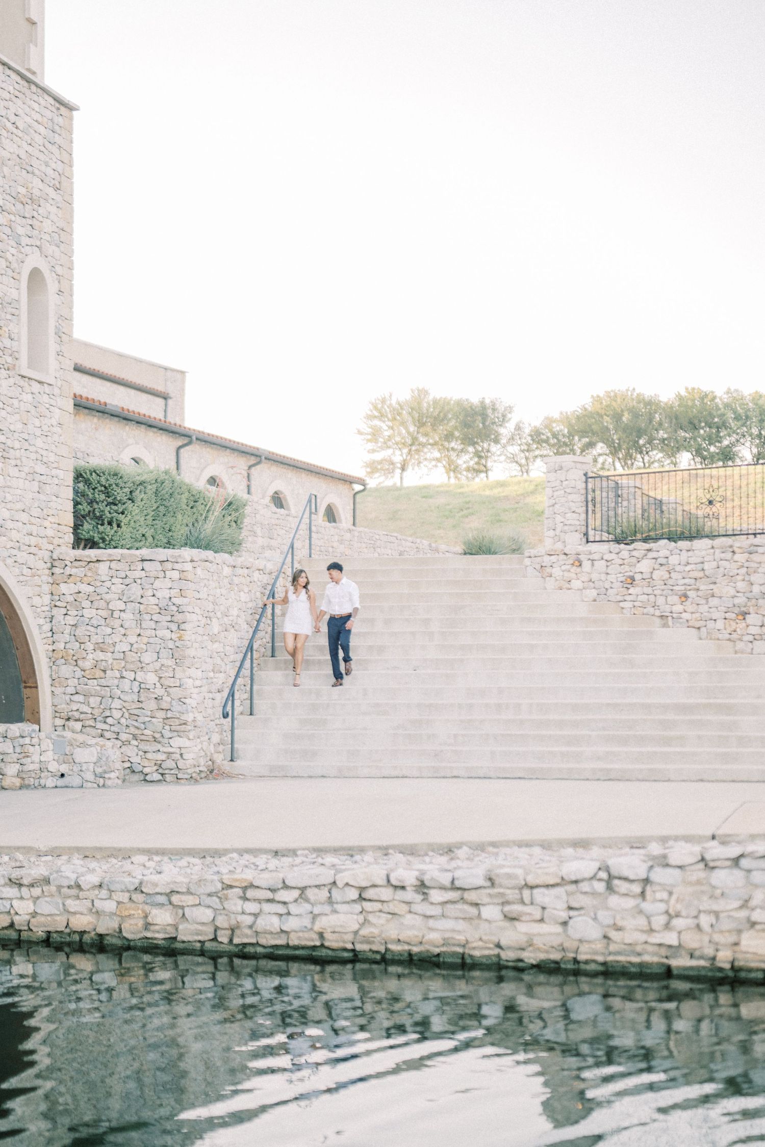Engaged couple holding hands walking down stone steps near a reflective pool at a scenic venue.