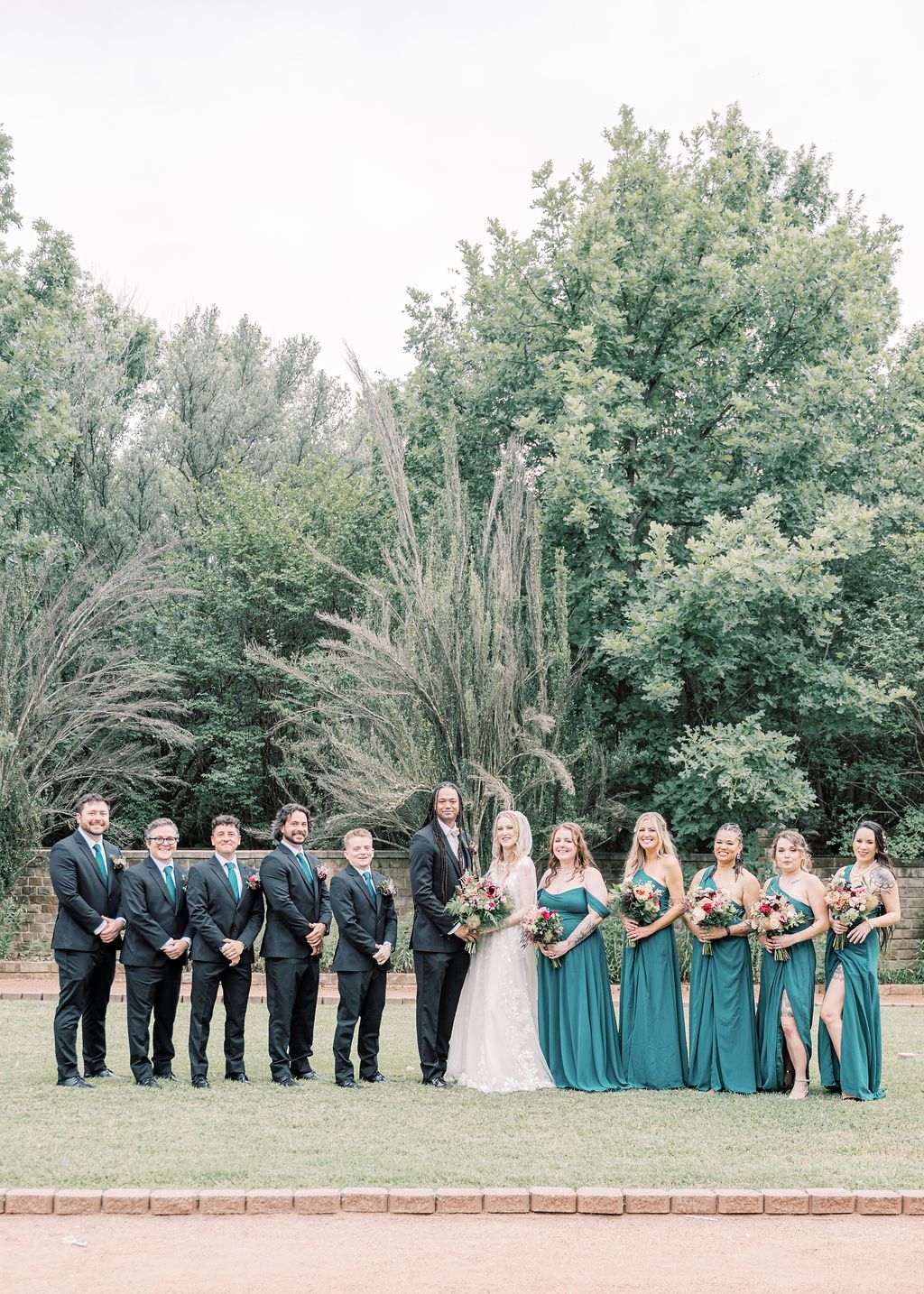 Wedding party posing outdoors, groomsmen in black suits and bridesmaids in teal gowns flanking bride and groom before lush green trees.