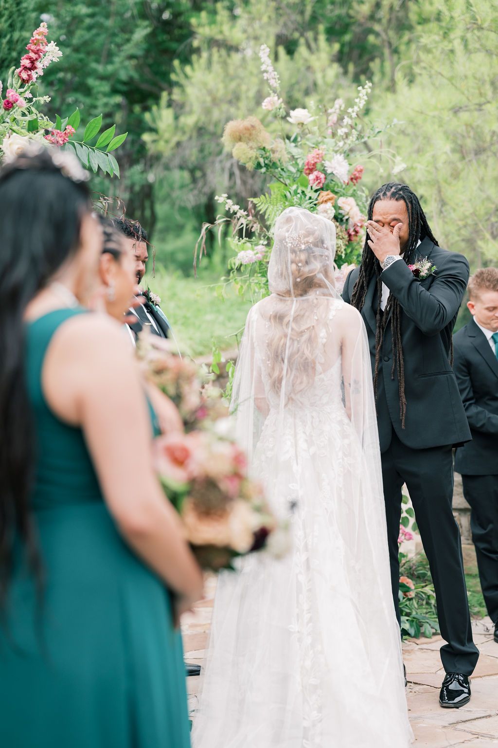 Emotional groom covers mouth in tears as bride in lace cape veil approaches floral altar outdoors.