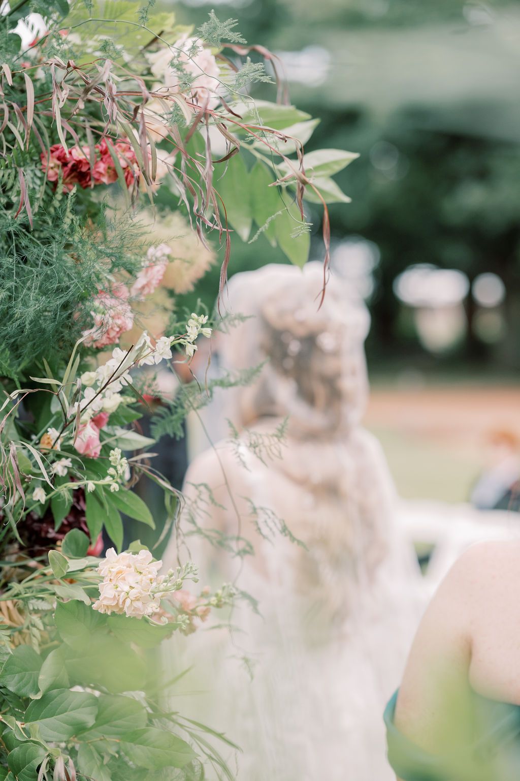 Lush floral wedding arch with greenery, roses, and trailing vines in soft focus with bride in background.