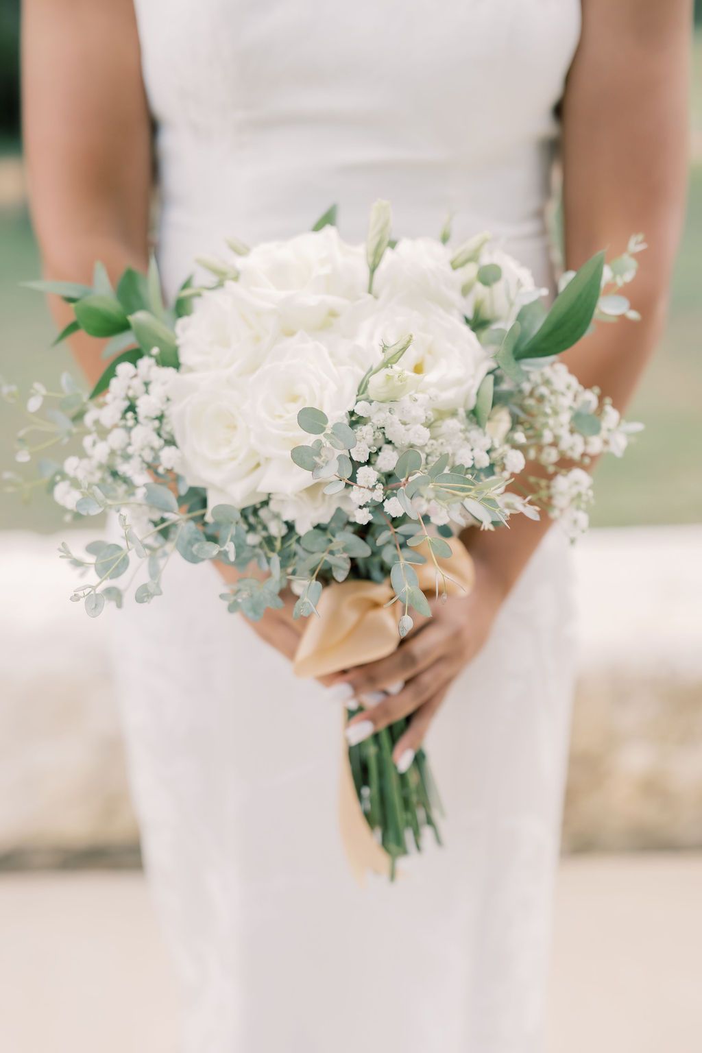 Bride in white dress holding elegant bridal bouquet of white roses, eucalyptus, and baby's breath tied with gold ribbon.