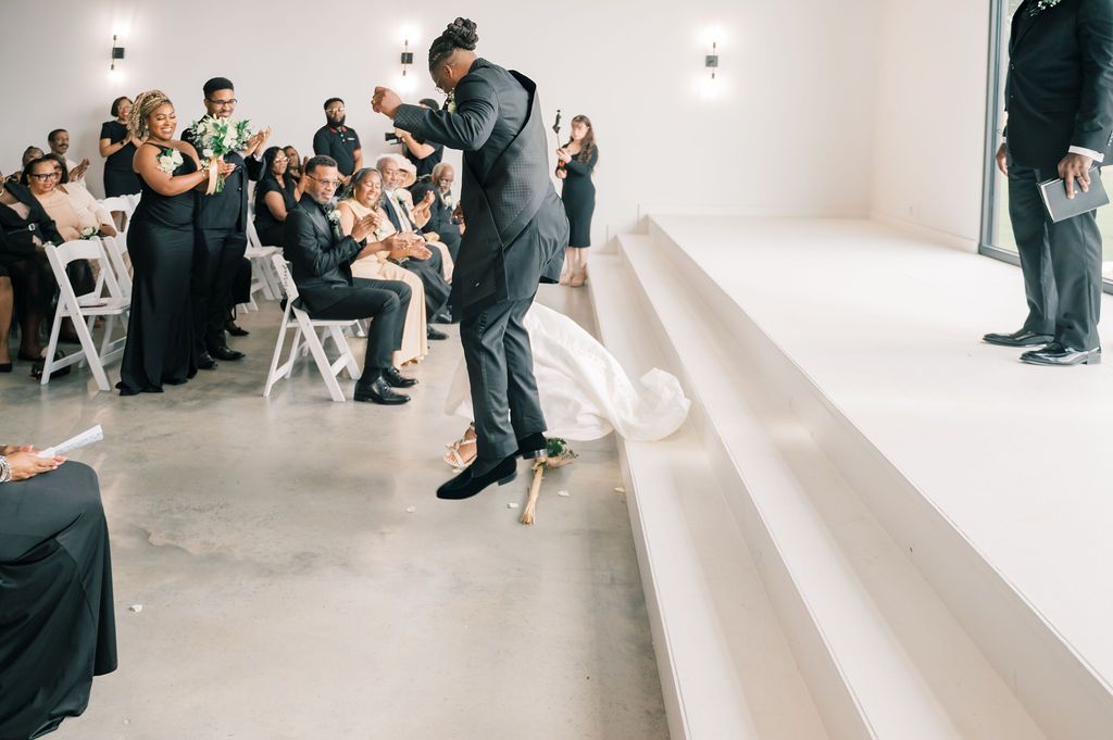 Groom leaps joyfully off stage steps during wedding ceremony as guests celebrate in background.