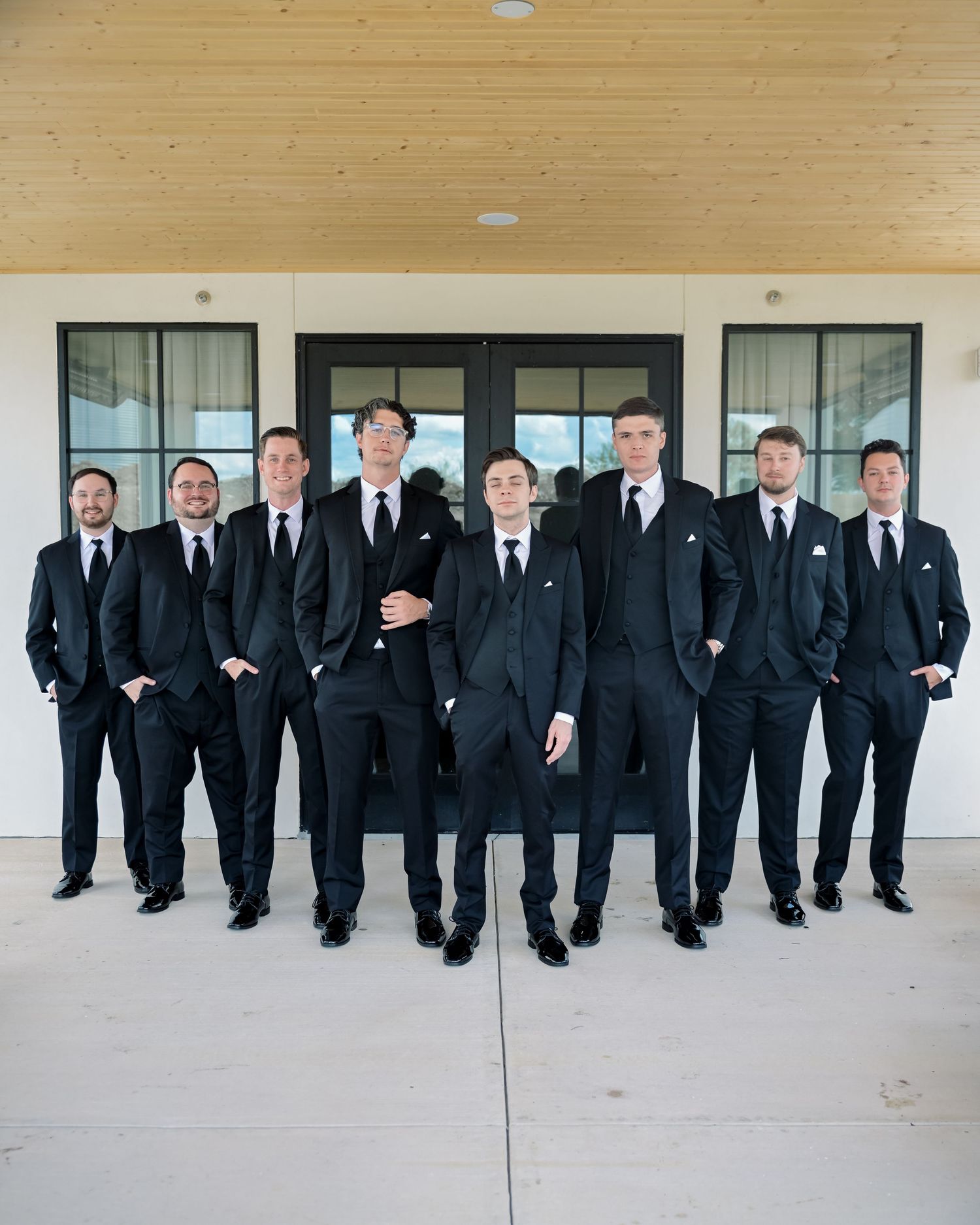 Nine groomsmen in matching black three-piece suits posing together outside a modern white wedding venue.