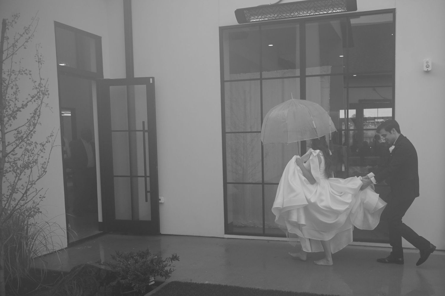 Groom shelters bride with umbrella while carrying her gown in black and white photo.