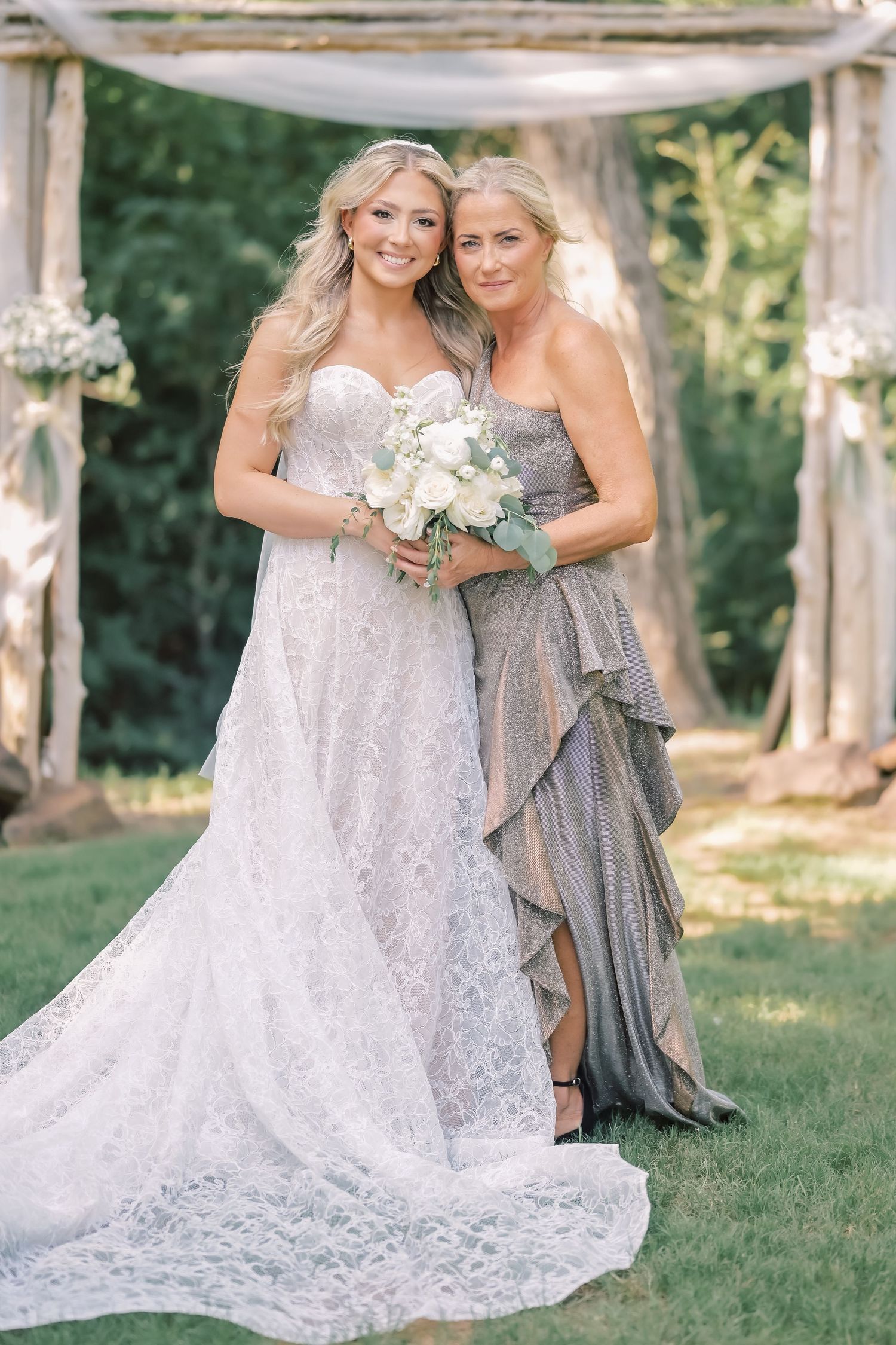 Radiant bride in lace gown holds white bouquet posing with mother in silver dress at outdoor wedding.