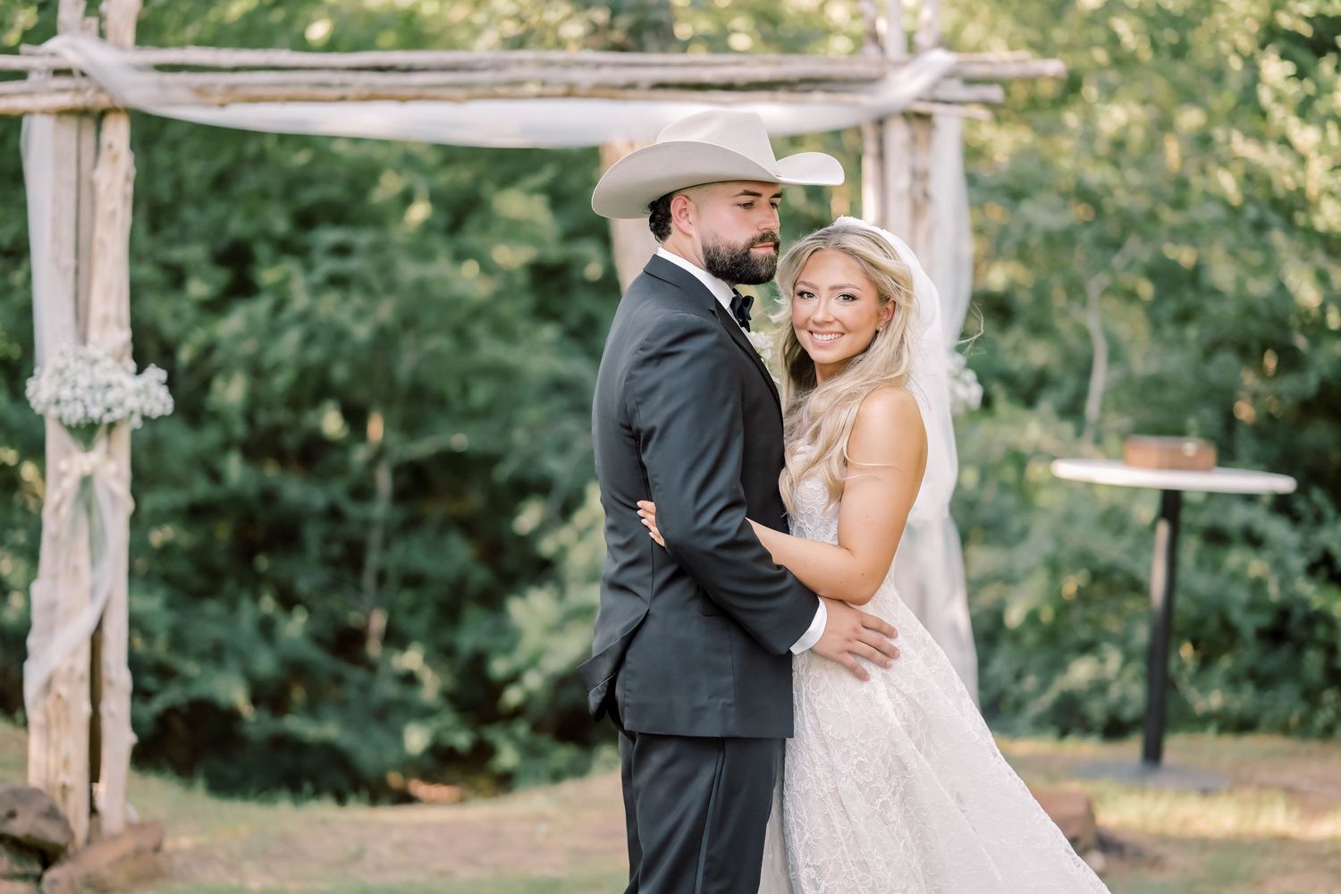 Bride and groom embrace under rustic wooden arch surrounded by lush greenery at outdoor ceremony.