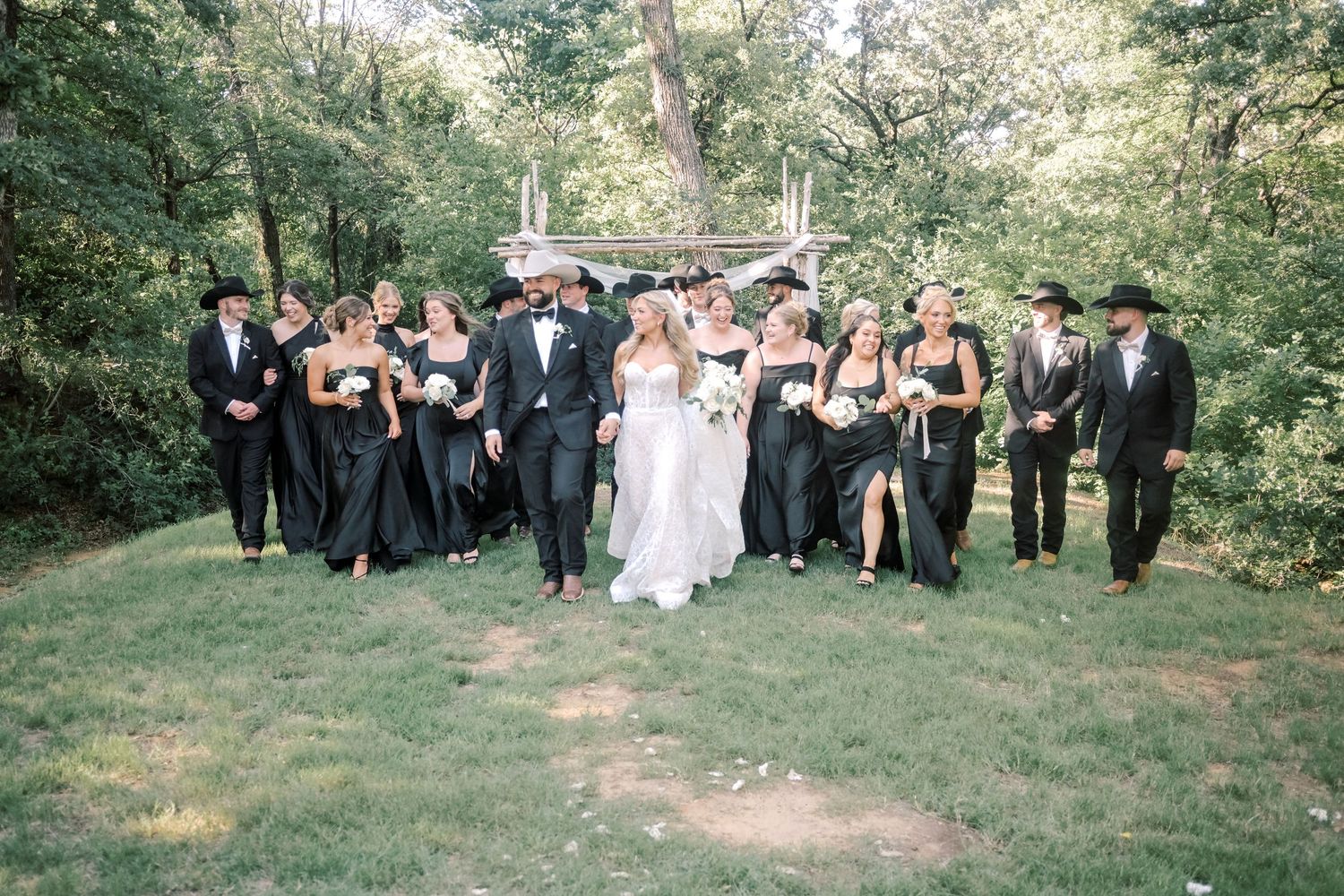 Wedding party in black attire and cowboy hats walk together on green lawn at outdoor ceremony.