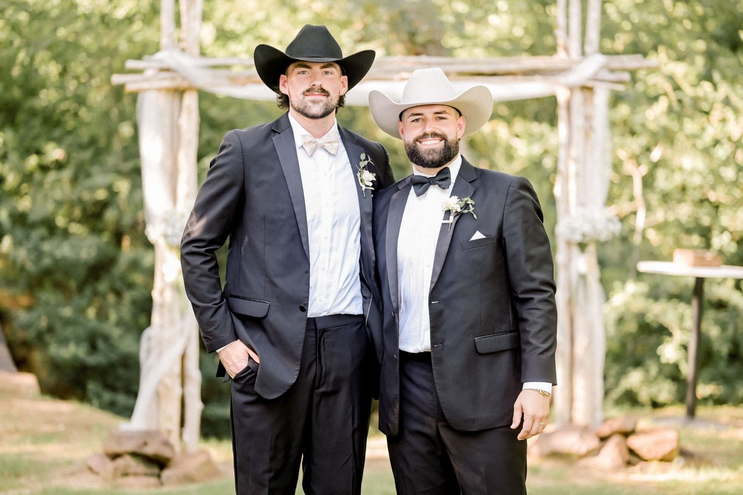 Two men in dark suits and cowboy hats posing together outdoors at a wedding ceremony.