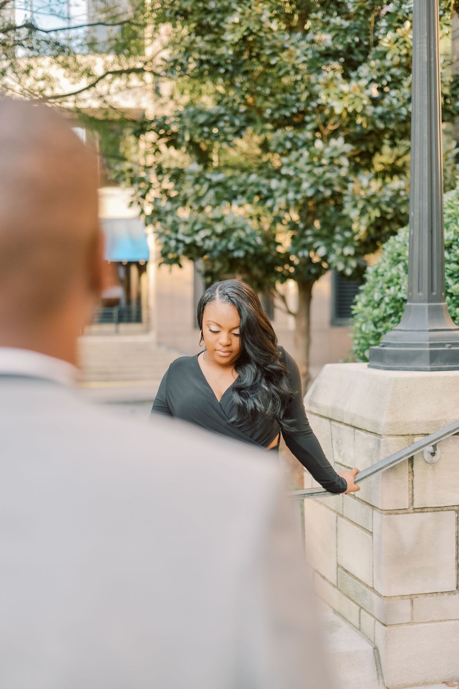 Woman in black wrap dress holds staircase railing as man in gray suit looks on in urban setting.