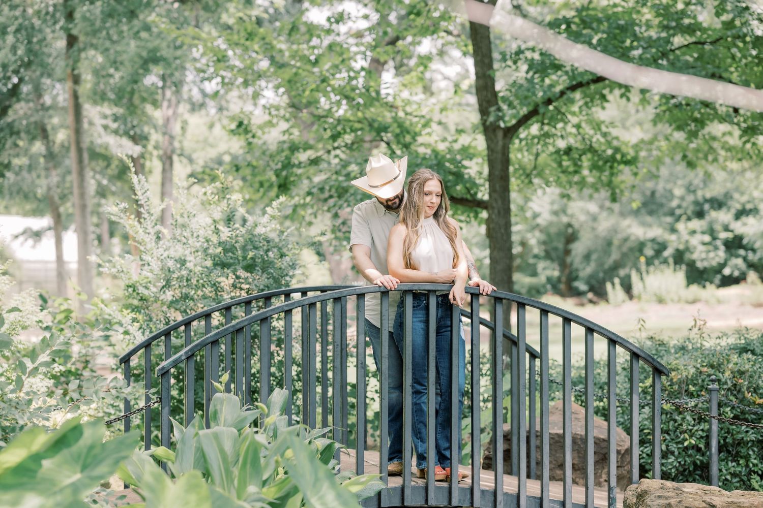 Couple standing on a curved iron bridge surrounded by lush green trees in a romantic outdoor engagement photo.
