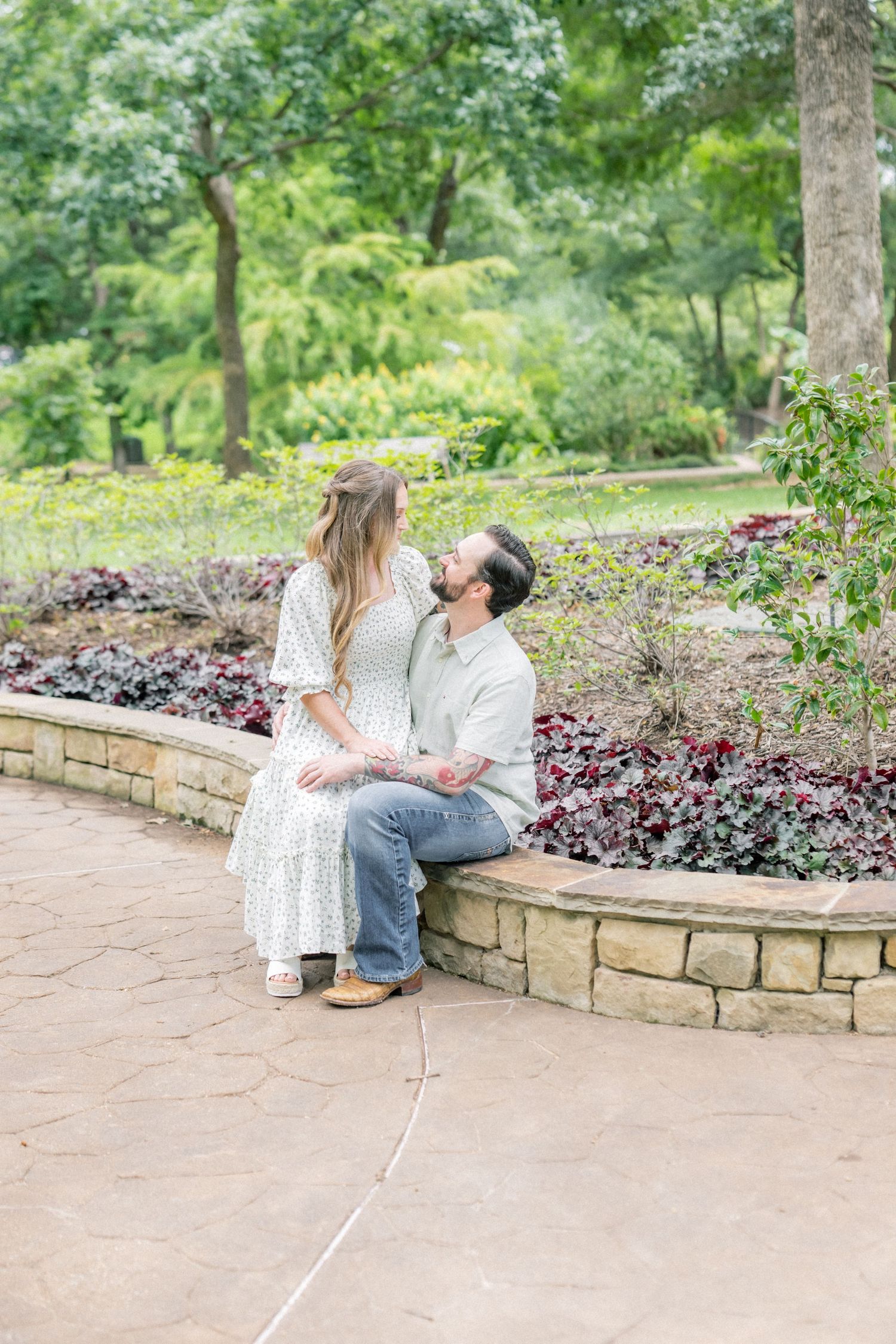 Couple sitting together on a stone garden ledge surrounded by colorful foliage and trees.