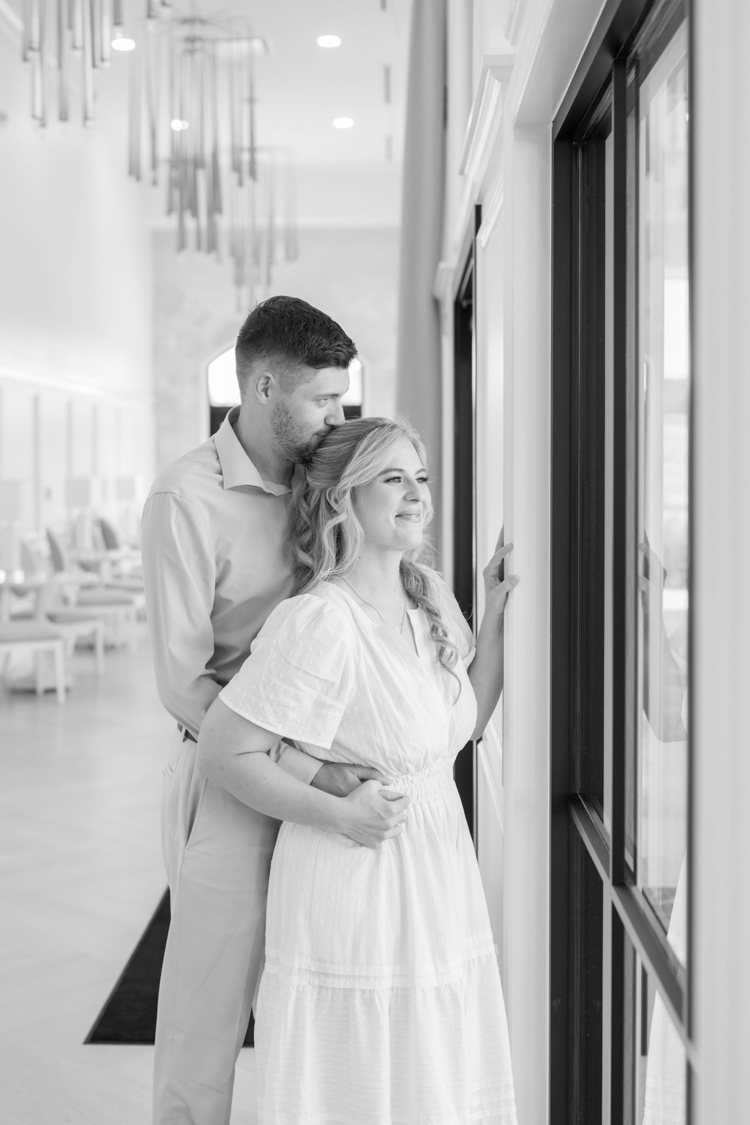 Black and white photo of a couple embracing by large windows inside an elegant venue with chandeliers.