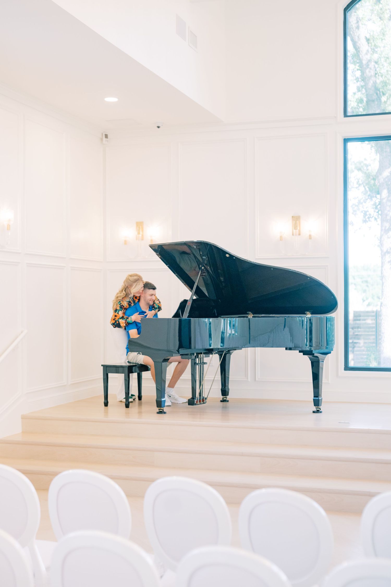 A couple sits together at a grand black piano in a bright, elegant white chapel interior.