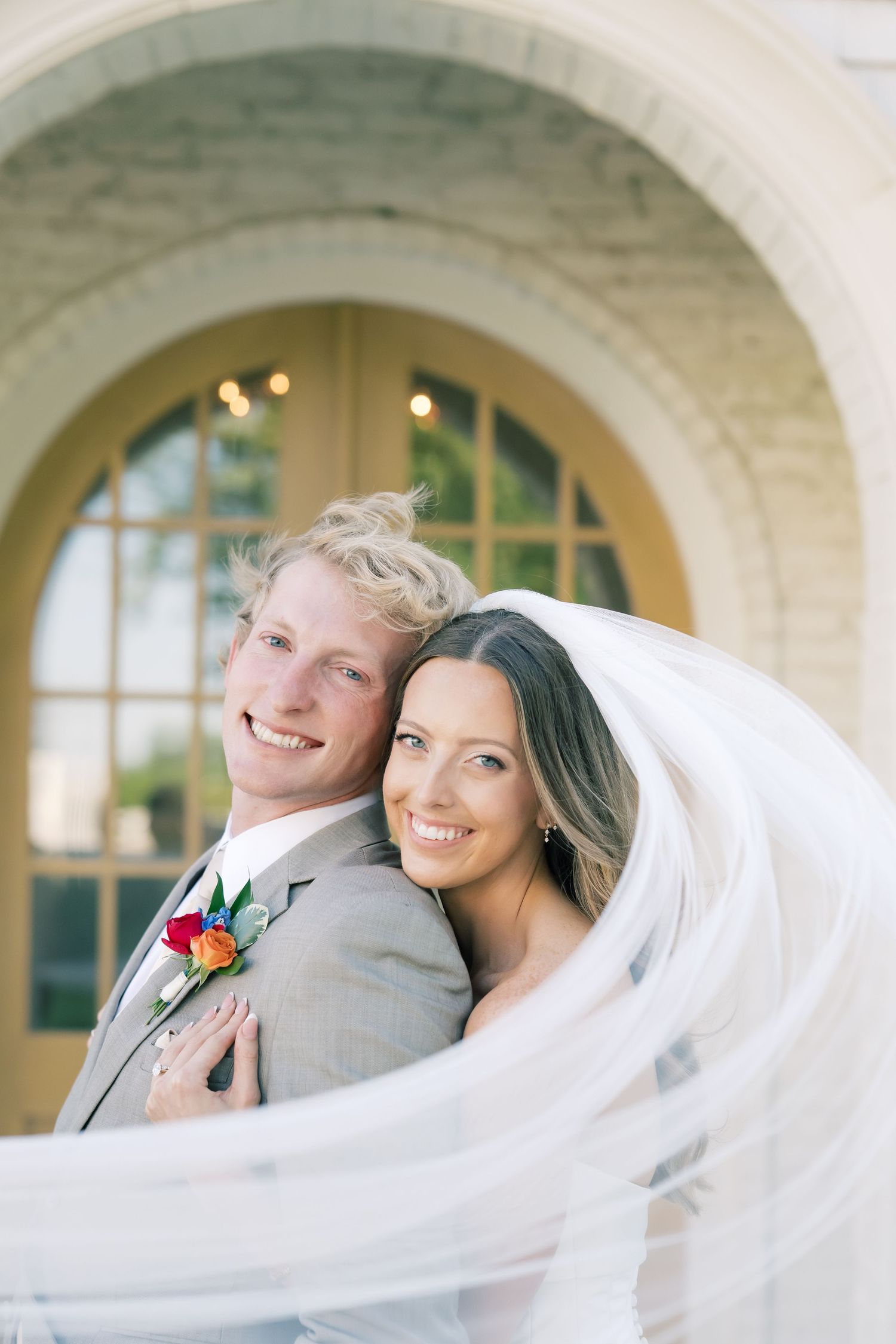 Smiling bride and groom pose together under an arched doorway, her veil flowing, he wears a gray suit with colorful boutonniere.