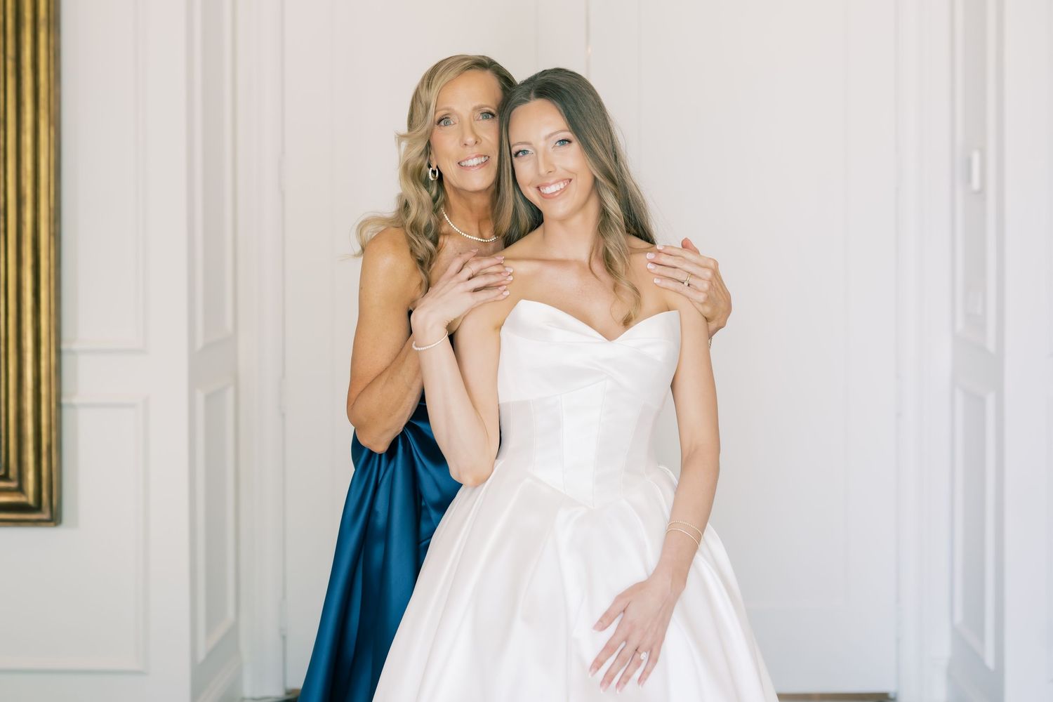 Bride in white strapless ball gown poses with woman in blue dress, both smiling in elegant white room.