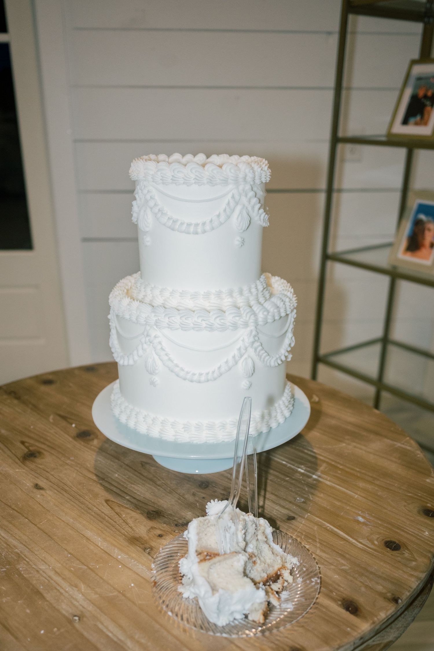 Elegant two-tier white wedding cake with pearl and swag details on a wooden table, with a slice served on a glass plate.