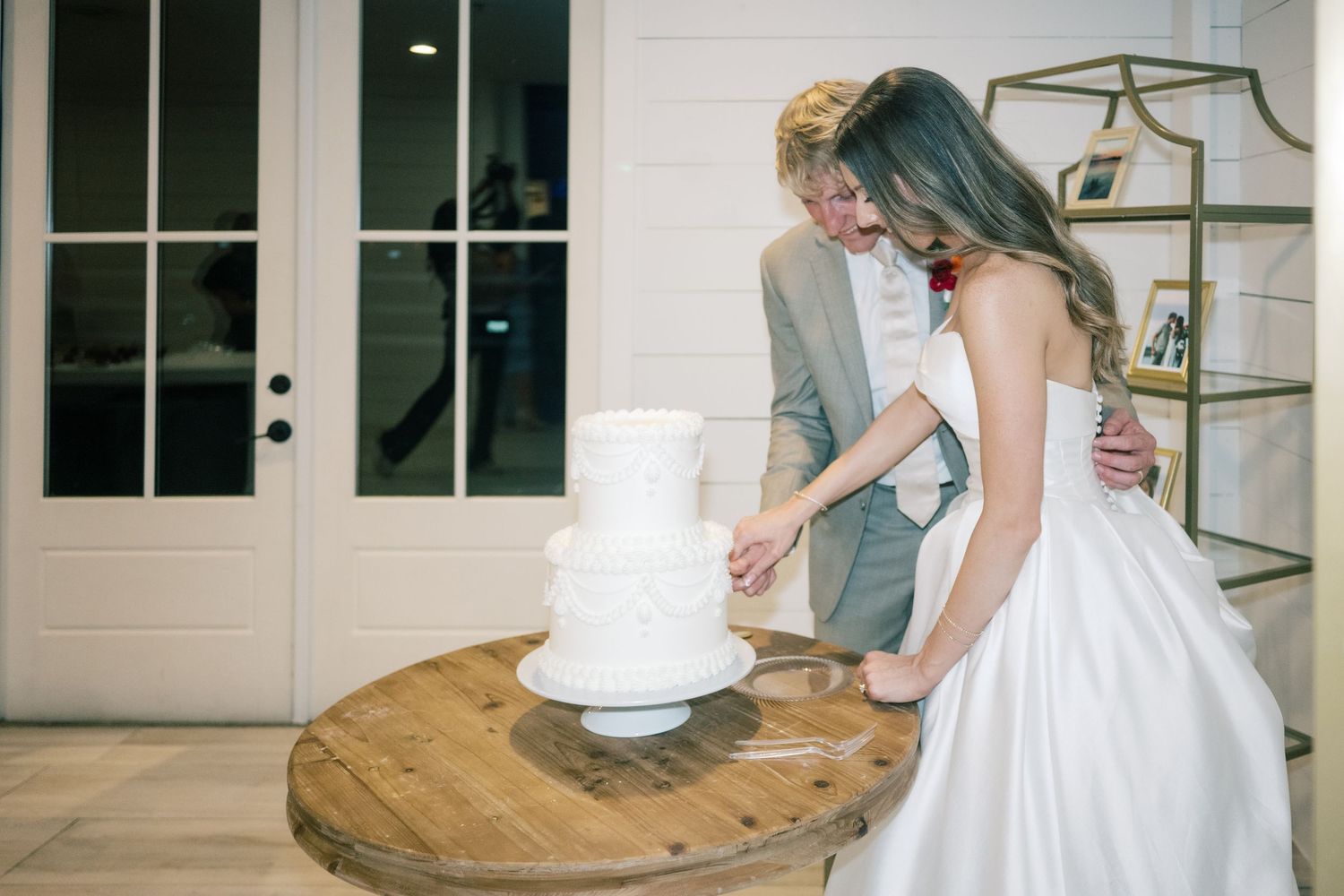 Bride and groom cutting white three-tier wedding cake on rustic wooden spool table at elegant indoor venue.