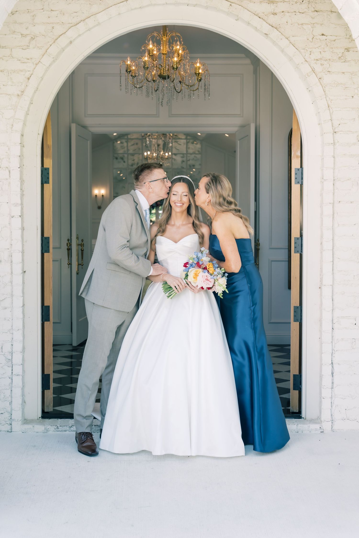 Bride smiling as father kisses her cheek and bridesmaid embraces her in elegant arched doorway with chandelier.